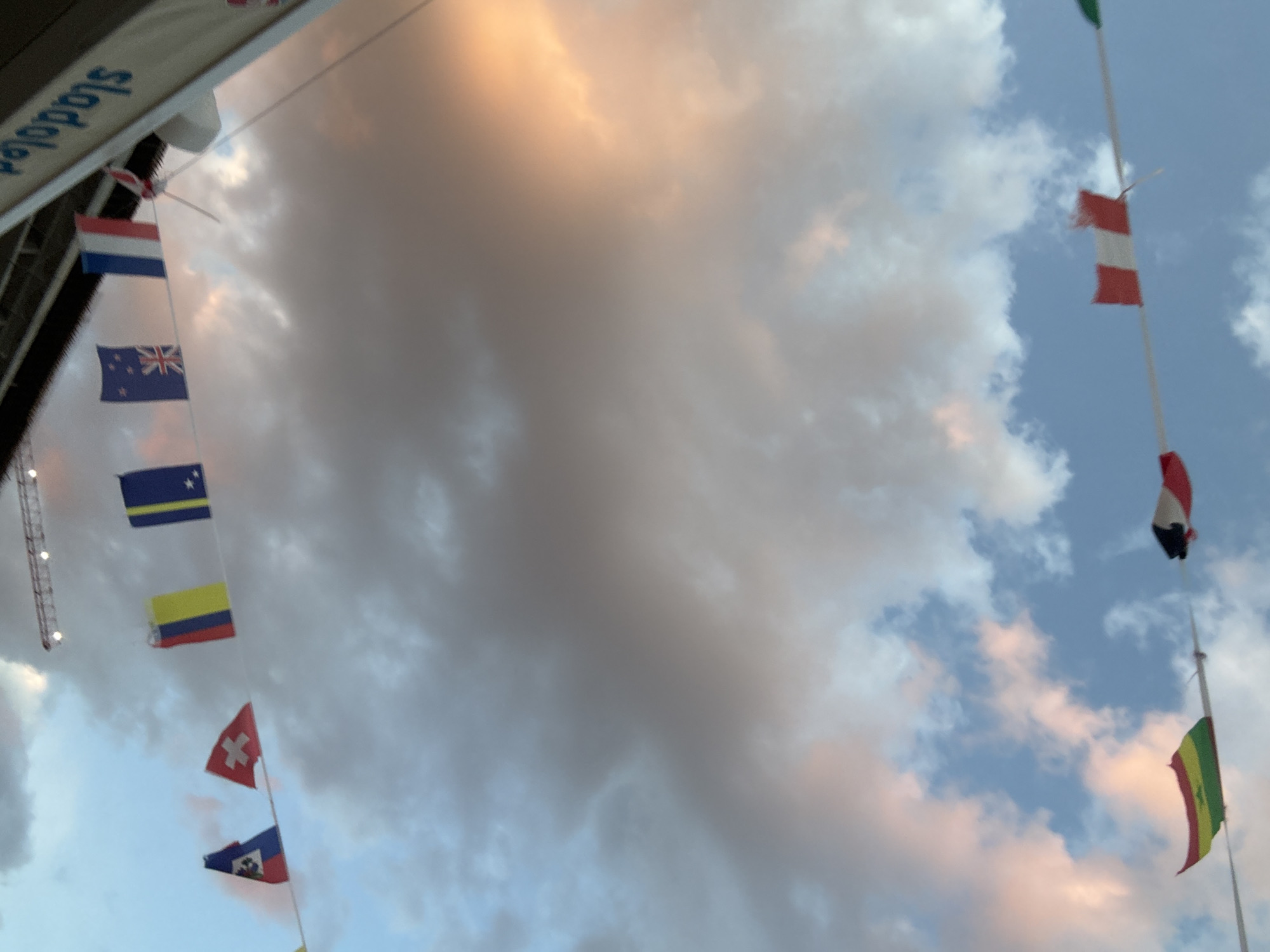 The image shows a sky with cumulus clouds in varying shades of grey, white, and with some orange hints. There are several national flags on poles visible. Some are: the flag of the Netherlands, New Zealand, Curacao, Columbia, Switzerland, and Haiti. On the right of the image, there is the flag of Austria and Bolivia. The location is likely Budva, Montenegro, due to the signs written in Montenegrin language. There is a tower crane visible. The flags are arranged linearly, which suggests they are hung for an event.