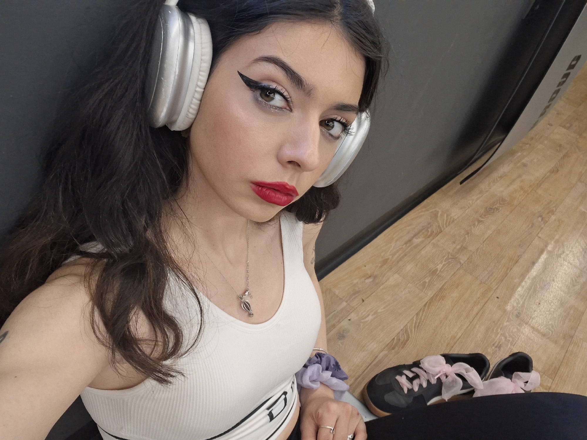 A young woman, likely in Sofia, Bulgaria, is captured in a close-up, high-angle selfie indoors. She has dark, wavy hair, fair skin, and is wearing prominent winged black eyeliner, red lipstick, and subtle blush. Large, silver over-ear headphones cover her ears. She is dressed in a white ribbed tank top, featuring a dark band with indistinct text below the bust. A silver necklace with a hot air balloon-shaped pendant hangs around her neck. On her left wrist, a light purple scrunchie is visible, and she wears a silver ring on her left ring finger.
The woman gazes directly at the camera with a serious or contemplative expression. She is leaning against a plain, dark gray wall or partition. To her right, a light wooden floor is visible, and next to her, a pair of black sneakers with distinctive pink ribbon laces are placed on the floor. Part of a larger dark gray structure or machine is visible behind her, with partial text appearing to spell "ANDUD" or similar. The setting suggests an indoor recreational space, possibly a gym or studio, where she might be taking a break from an activity or simply posing for a photo.
