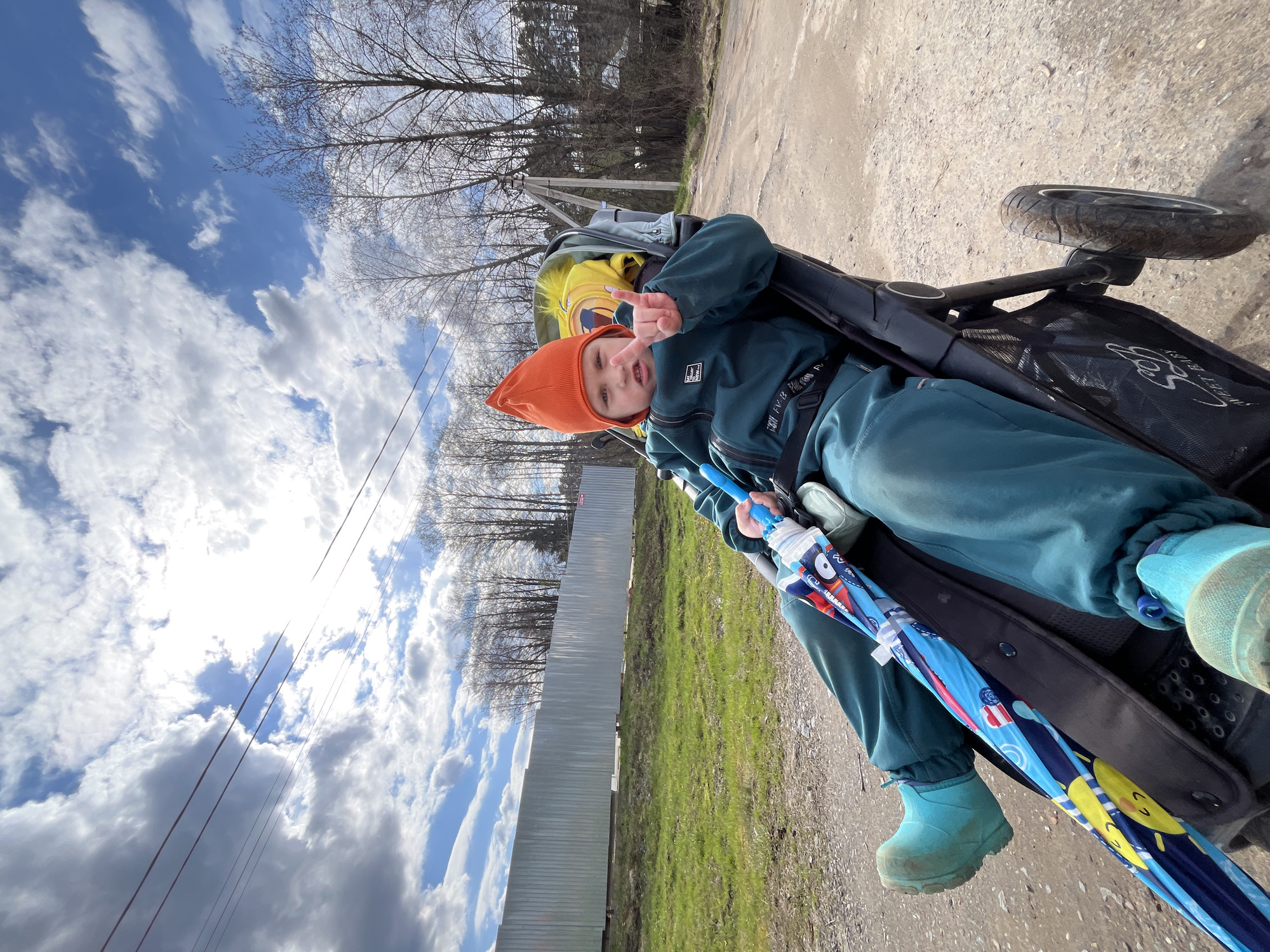 This tilted outdoor photograph, taken in Chaikovsky, Russia, captures a young child seated in a dark-colored stroller on a dirt or gravel path. The child wears an orange knit hat, a teal jacket, and matching pants, with light blue boots. They are looking directly at the camera, making a peace sign with their right hand, and holding a closed blue umbrella featuring cartoon designs, including a sun.
The background features a bright blue sky with scattered white clouds, and a noticeable sun glare emanates from the upper right portion of the sky. Bare trees, characteristic of late winter or early spring, line the horizon. A tall, corrugated metal fence runs horizontally through the mid-ground, separating the path from patches of green grass. Power lines are also visible cutting across the sky.
The stroller has "Sweet Baby" text visible on its side mesh pocket. The child's jacket has some indistinct text on it, possibly "PLAY". The image conveys a cheerful and casual daytime outing.