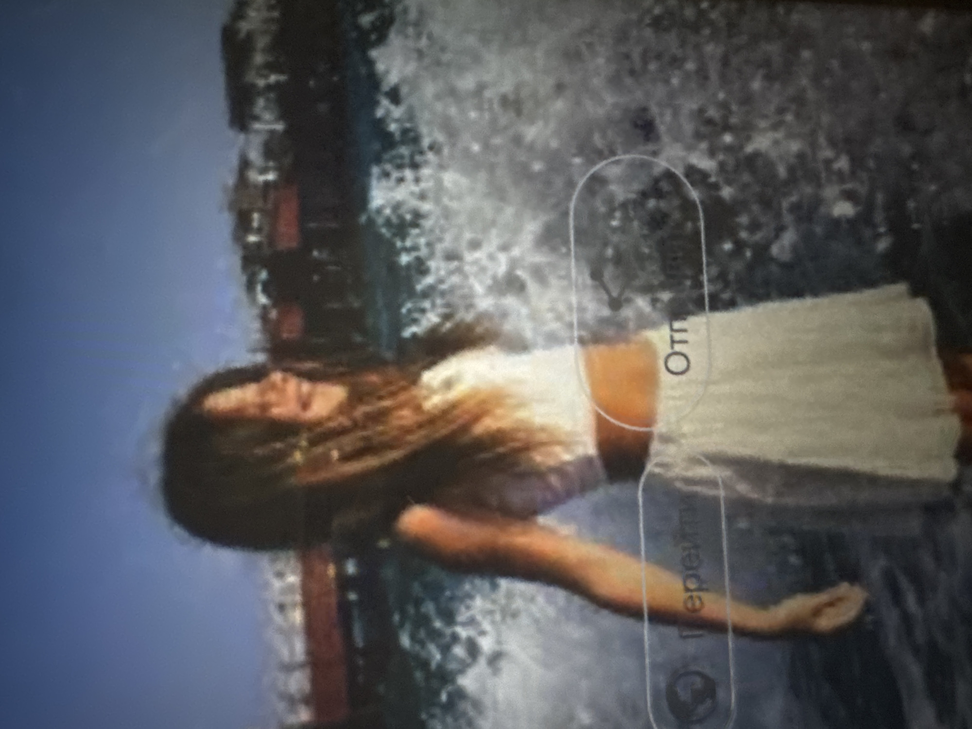 An image, rotated 90 degrees counter-clockwise, depicts a woman posing in front of a substantial water feature, likely a waterfall or large fountain. She has long, light brown hair, which appears somewhat wet and flows around her head. Her eyes are closed or cast downwards, and she has a gentle, serene expression. She is wearing a light-colored (possibly white or off-white) strapless top and a flowing, light-colored skirt.
The background primarily consists of a large volume of white, splashing water. To the upper left, a clear, bright blue sky is visible, indicating an outdoor, daytime setting with good weather. Darker, possibly stone or concrete structures are partially visible behind the water, suggesting a constructed water feature.
The image itself appears to be a photograph of a screen, exhibiting some blurriness and visual artifacts consistent with screen capture. An overlay from a device interface is visible, featuring a vertical translucent rectangle. Within this rectangle, Cyrillic text is discernable: "Отп" (Otp) at the top and "Перейти" (Pereyti), meaning "Go" or "Proceed," at the bottom. Below "Перейти," there is a small icon, possibly related to sharing or linking.