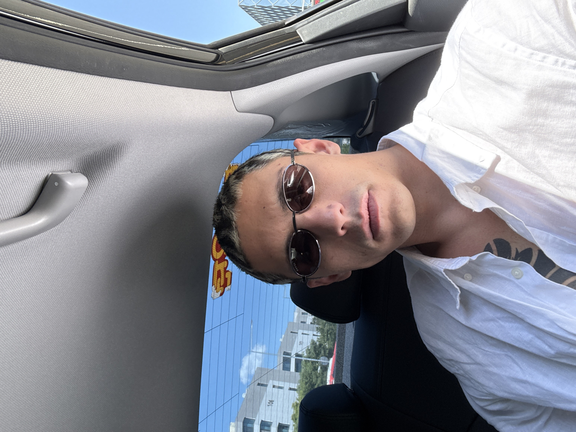 A man in a white shirt and sunglasses is seated in a car with an open sunroof. Through the rear window, buildings are visible in a cityscape. The Gucci sticker on the rear windshield suggests the location is in Seoul, South Korea.