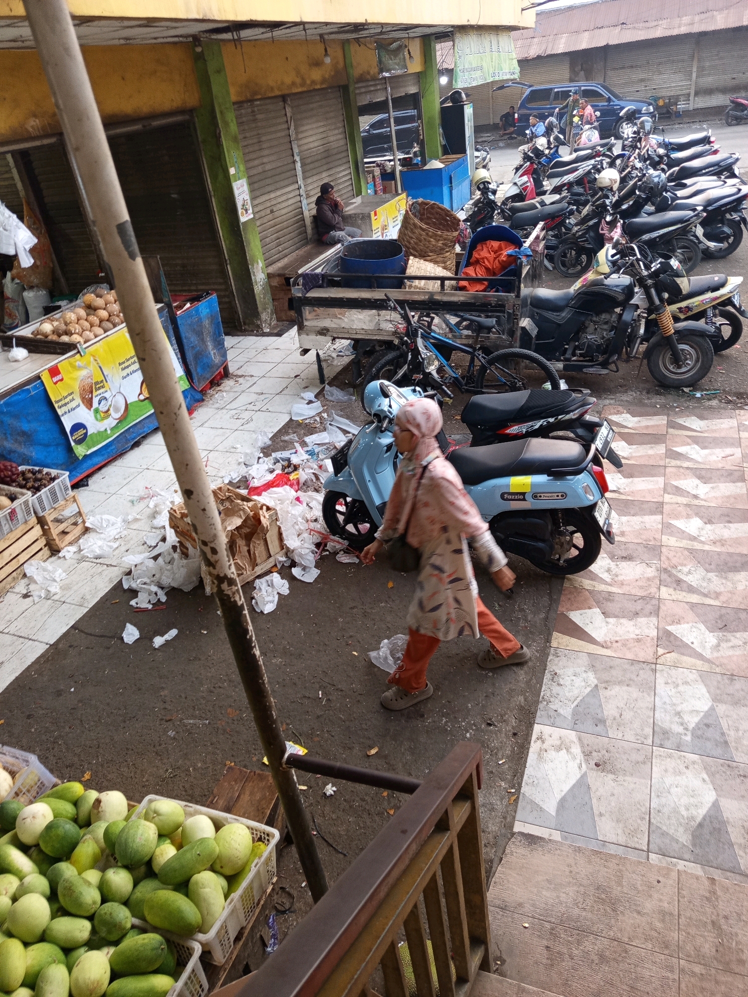 This image depicts a street market scene in Karawang, Indonesia. In the foreground, a person wearing a hijab and a light-colored patterned tunic with orange pants walks across a tiled pathway. Scattered on the ground are discarded plastic bags and other debris. To the left, a basket overflowing with green fruits, possibly cucumbers or melons, sits on a wooden crate. Further back, a light blue scooter is parked near a pile of brown cardboard boxes and more scattered waste. Behind the scooter, numerous motorcycles are parked in rows, and a blue SUV is visible. The market stalls have metal roller shutters, some open and some closed. A person is seated near an open stall in the background, smoking. The overall environment suggests a busy, somewhat untidy market area.