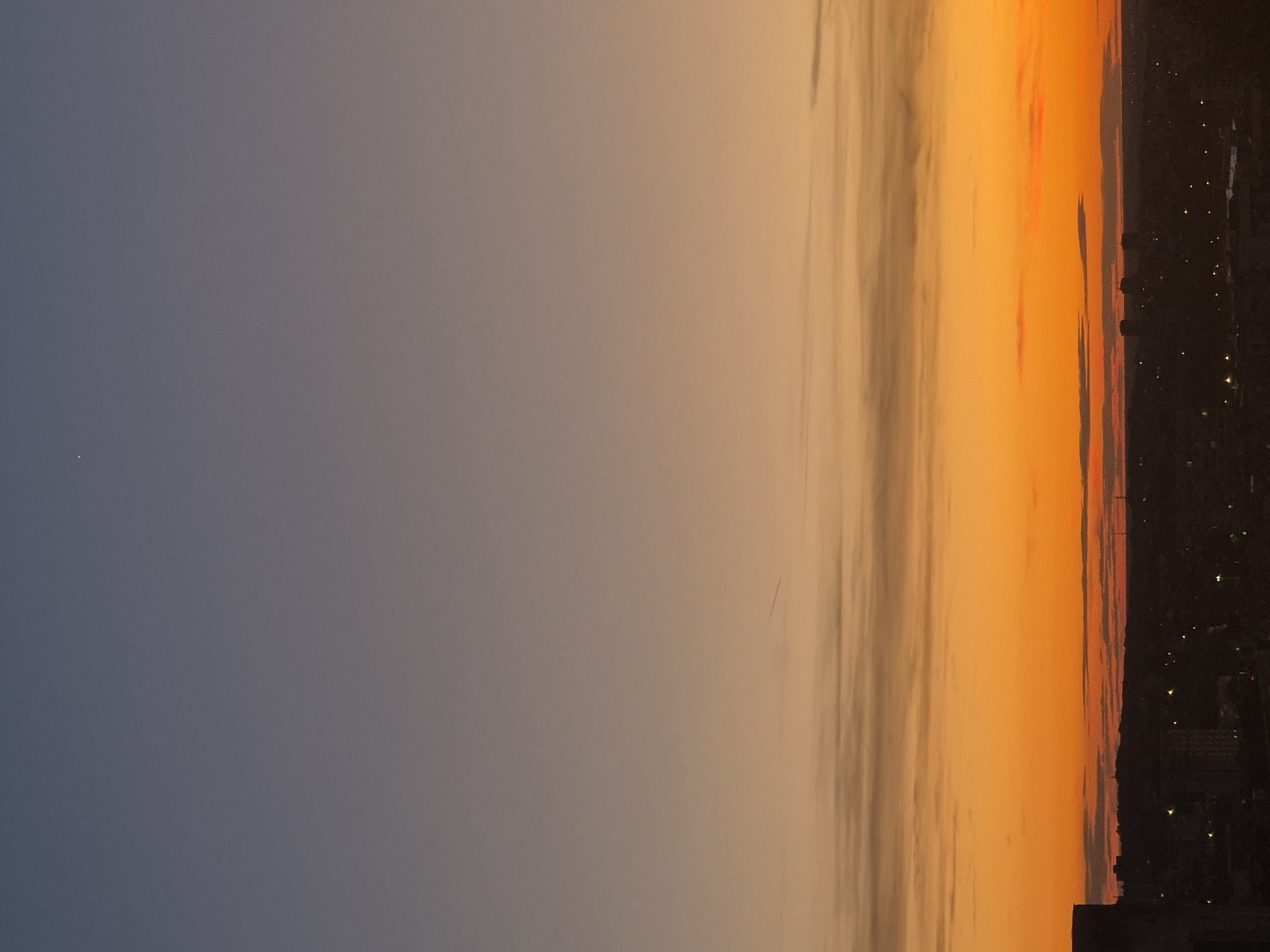 The image depicts a sunset over a city skyline. The sky transitions from a deep blue at the top to a vibrant orange and yellow near the horizon. A few wispy clouds are visible, stretched horizontally across the sky. There is a faint contrail from an aircraft in the upper middle section of the sky, and a small, bright celestial body is visible in the upper left corner, possibly a planet or a bright star. The cityscape at the bottom is silhouetted against the colorful sky, with buildings and lights indicating an urban environment. The overall scene is one of twilight in a city.