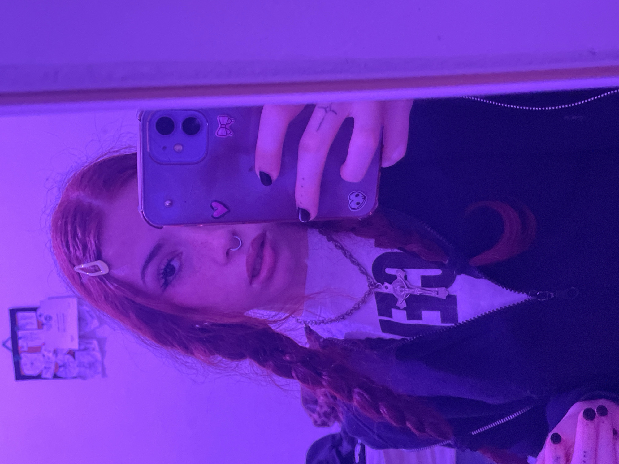 A person with red hair in braids, a nose ring, and black nail polish is taking a selfie in a mirror using a cell phone. The phone has stickers on the case. The person is wearing a black jacket over a white shirt with a large, dark graphic. A silver necklace with a cross pendant is also visible. The reflection shows a small white hair clip in the person's hair and a cluttered shelf on the wall behind. The lighting is predominantly purple. There is no indication of the city of Buenos Aires, Argentina, or any location.