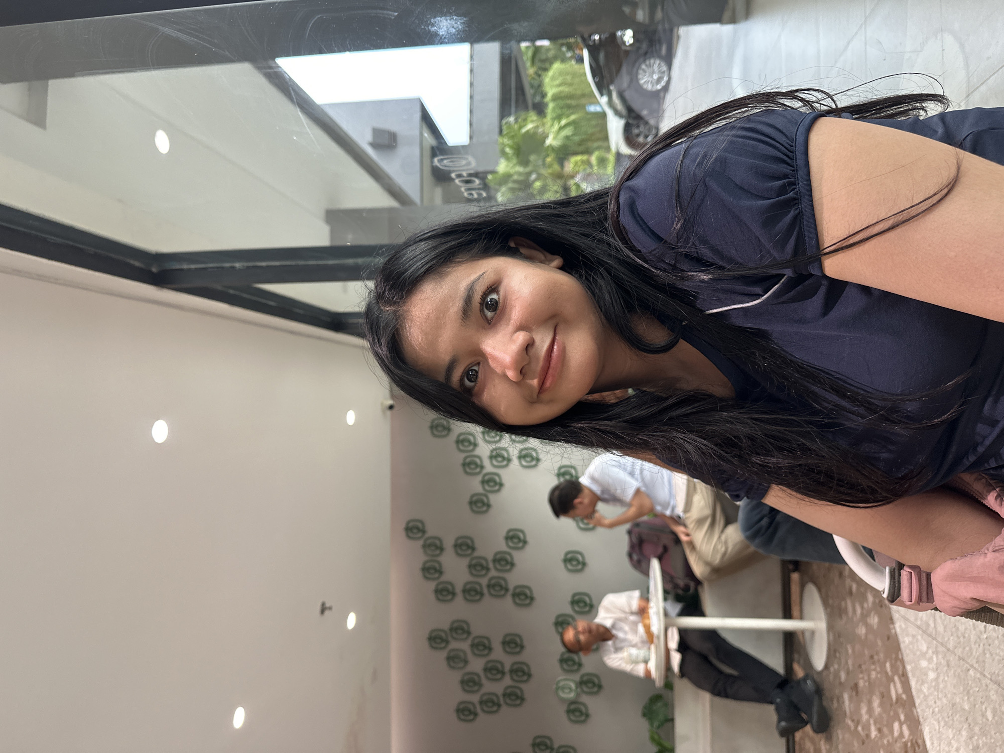 The image shows a woman with long black hair, wearing a dark blue t-shirt. She is positioned in the foreground, looking directly at the camera. Behind her, through a glass window, are other people. One man is seated at a table, another man is standing. The interior space features a white wall with a pattern of small, green circular designs. The location is a cafe in North Jakarta, Indonesia.