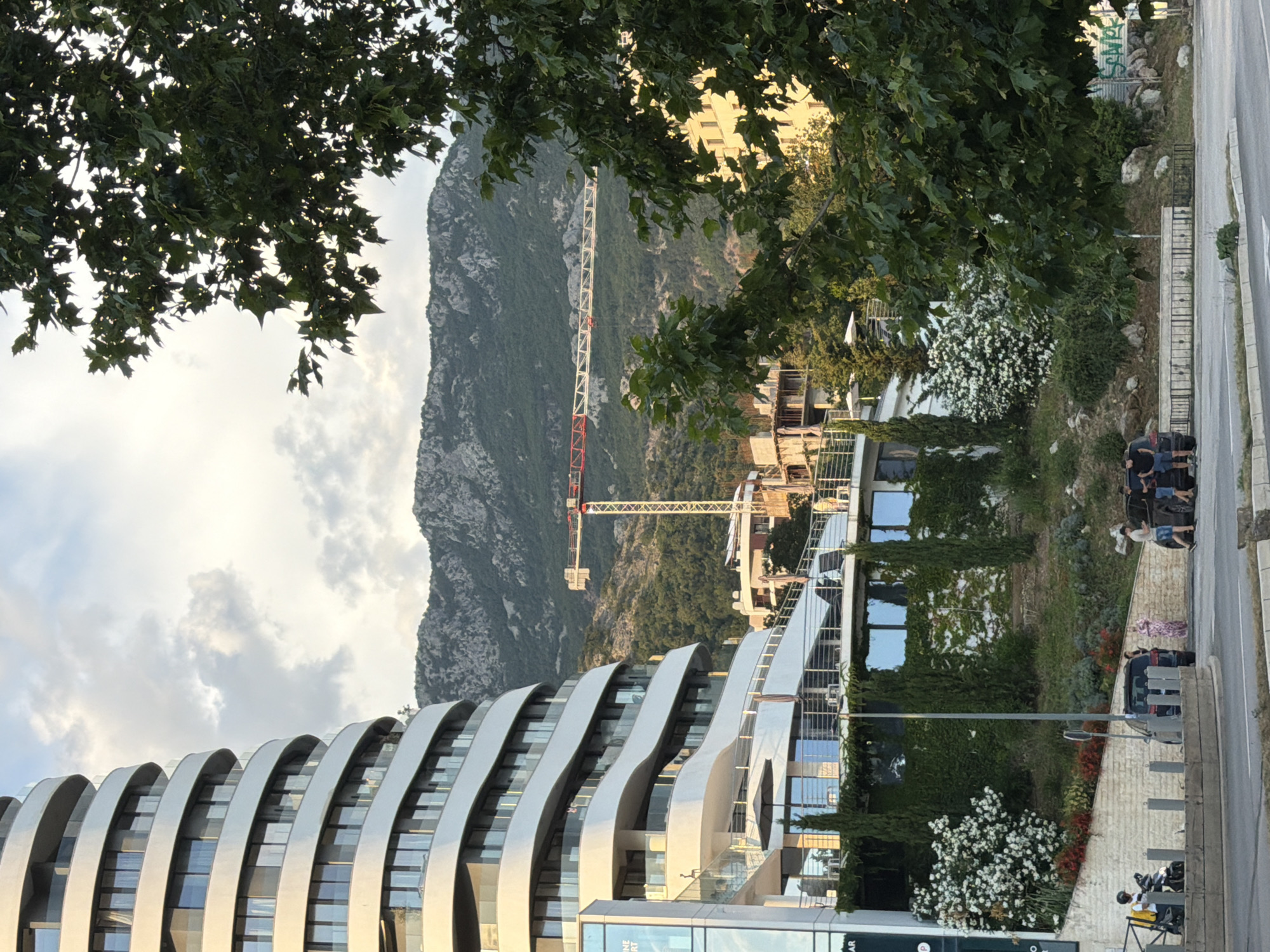 The image depicts an outdoor scene with a modern, curved building in the foreground, featuring numerous reflective windows and sleek, silver architectural elements. Lush green trees with dark leaves overhang the top of the frame. In the background, a towering construction crane with red and white sections is visible against a rugged, green mountainside. The sky is partly cloudy, suggesting it might be late afternoon or early evening.
Several people are visible in the image. Near the right side, a group of individuals, some standing and others sitting, are gathered near parked vehicles. One person is seated on a motorcycle in the lower right corner. Another person is seen in the middle ground near the building, and a woman in a patterned dress is standing near a car.
The setting appears to be urban or a mixed urban-natural environment, with the contemporary building and a road contrasting with the natural mountain landscape. The presence of the construction crane indicates ongoing development in the area. The overall mood is serene, with the natural elements like trees and the mountain providing a backdrop to the man-made structures. There are no prominent text elements visible in the image, aside from what might be signage on the building, which is too small to read.