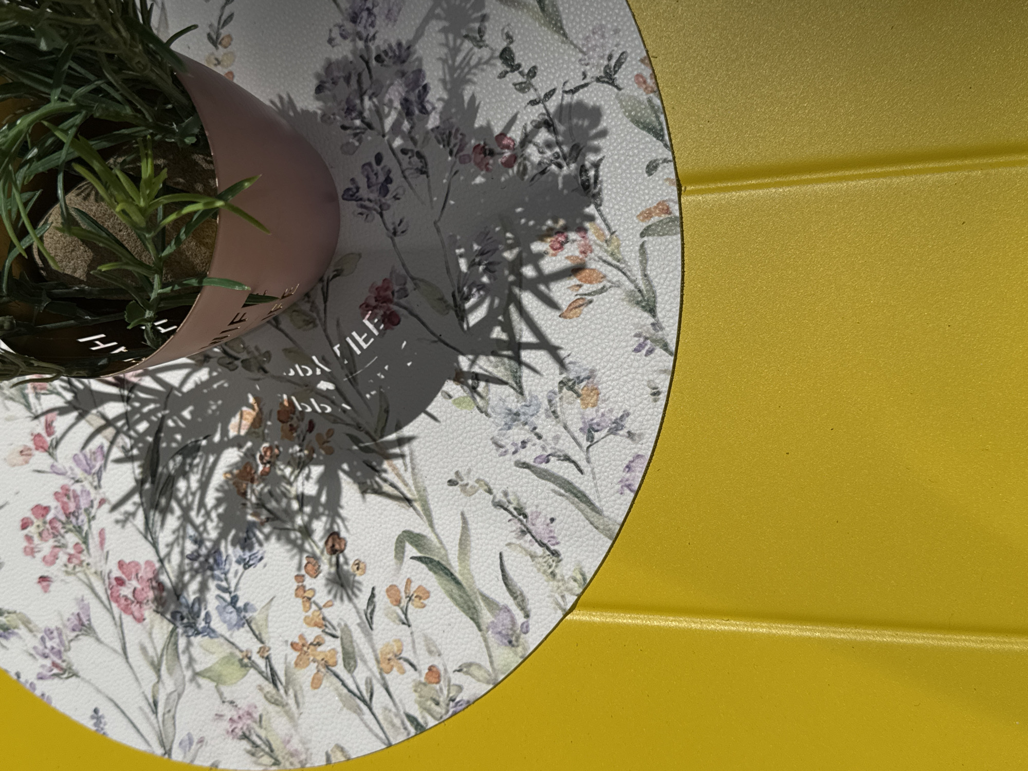 A high-angle shot reveals a yellow, horizontally-grooved surface covering a portion of the frame. On the left side, a circular floral-patterned mat is partially visible, partially covered by the shadow of foliage from a plant in a light-pink metal pot. The pot contains green plants. The overall light appears to be natural daylight.