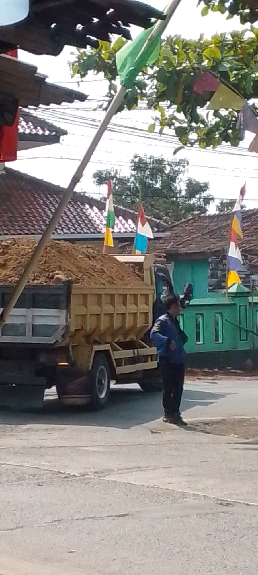 A man in a blue jacket and black pants stands on the right side of the frame, looking to the left. He is positioned to the right of a yellow dump truck loaded with dirt. The truck is parked on an asphalt road. Behind the truck, a green building with windows is visible. Decorative flags in various colors are strung across the upper portion of the image, suggesting a festive or celebratory occasion. The background features several houses with red tiled roofs and lush green trees. The scene appears to be set in a residential or semi-urban area in Cilaku, Indonesia, possibly during daytime.