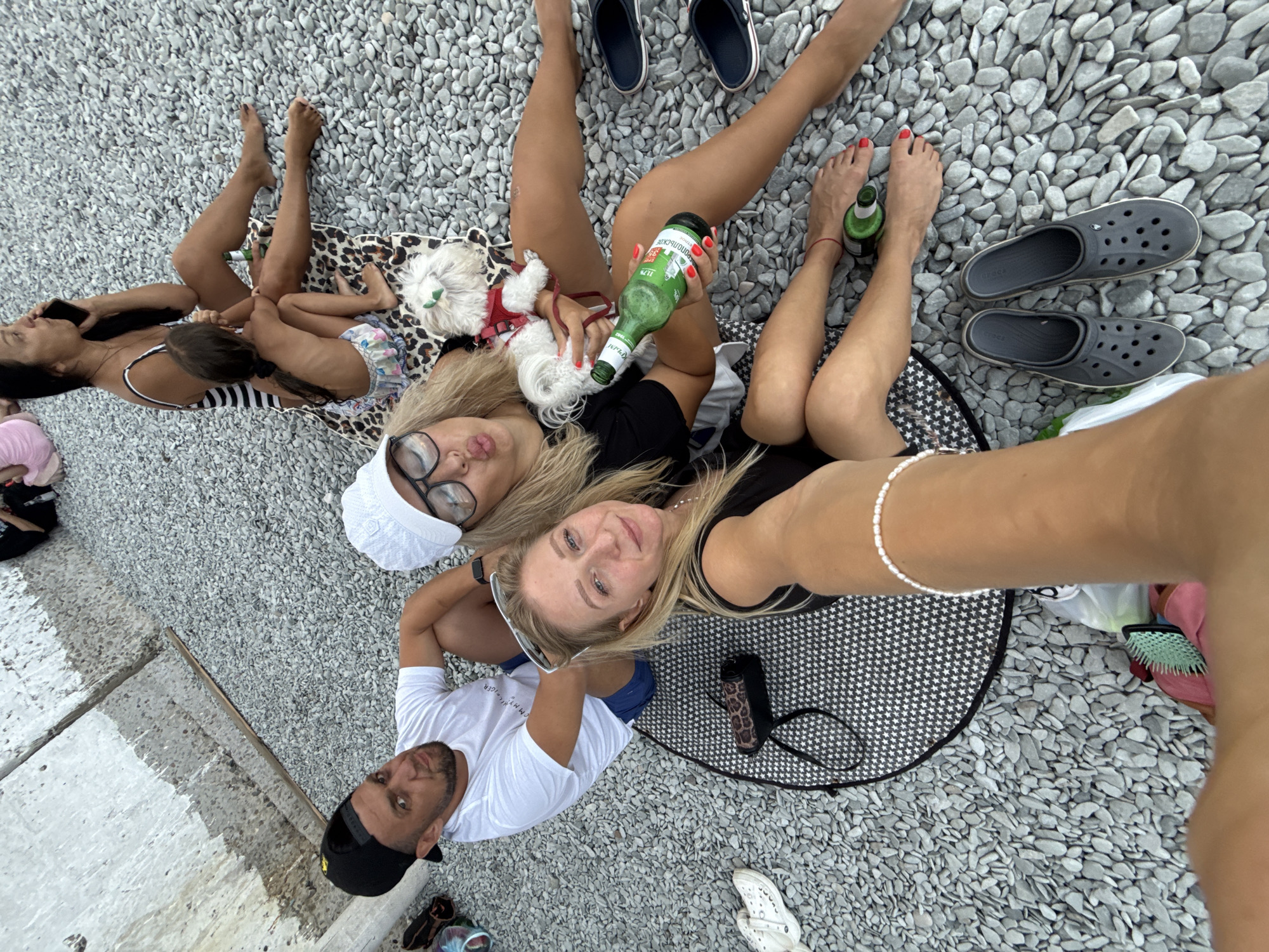 The image depicts a group of people and a dog relaxing on a pebbled beach. In the foreground, a woman with blonde hair, wearing sunglasses and a white cap, is taking a selfie. She is holding a green bottle of beer. Next to her is another woman with blonde hair, wearing a black top and a pearl bracelet, who is also looking towards the camera. A man with a black baseball cap and a white t-shirt is sitting beside them, looking directly at the camera. Further in the background, a woman in a striped swimsuit is talking on a mobile phone while holding a young child. A small white dog with a red harness is held by the woman in the swimsuit. The environment consists of a gray, pebbled beach with some concrete structures in the background. There are several pairs of shoes scattered around the scene, including Crocs and flip-flops. The overall atmosphere suggests a casual outing on a warm day. The location is identified as the city of Partenit, Ukraine.
