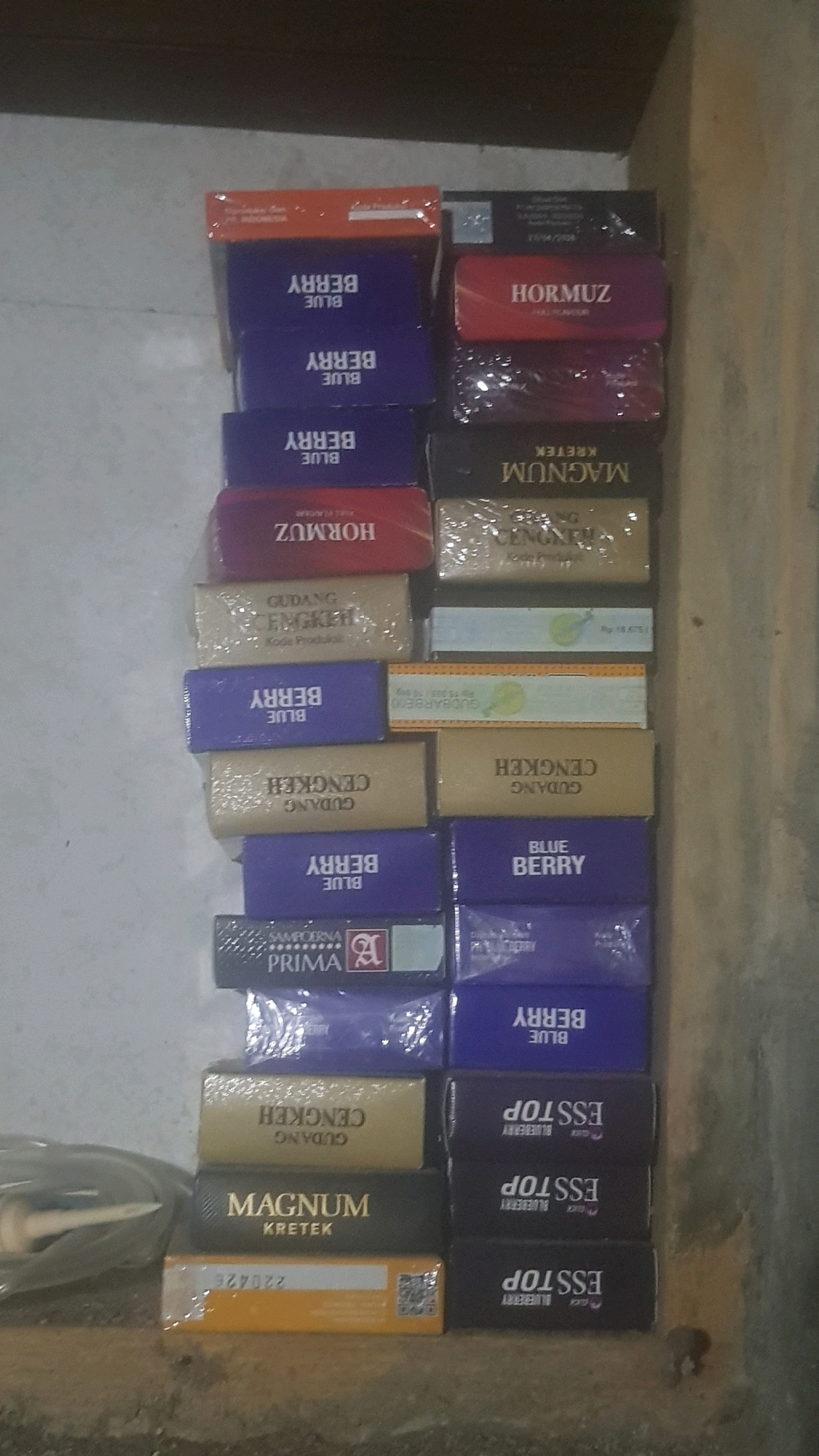The image shows a close-up, overhead view of a stack of cigarette packs on a shelf in a retail setting in Bakalan, Indonesia. The packs are arranged in two columns. The left column is taller, with packs of "BLUE BERRY," "HORMUZ," "GUDANG CENGKEH," and "MAGNUM KRETEK." The right column has packs of "HORMUZ," "MAGNUM KRETEK," "GUDANG CENGKEH," and "BLUE BERRY ESSSTOP." The lighting is dim and appears to be artificial. The background is blurred and indistinct, suggesting an indoor environment. There are no people or animals visible in the image.