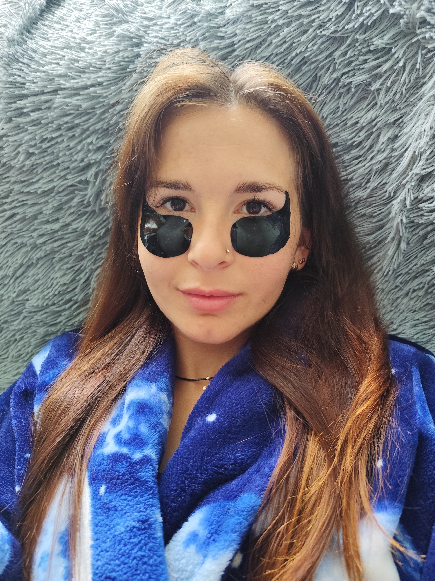 A young woman with long, brown hair, wearing a blue tie-dye bathrobe and black under-eye patches, is lying down on a soft, grey, fuzzy surface. She is looking directly at the camera, with a slight smile. Her hair is parted in the middle and falls over her shoulders. There is a thin necklace around her neck and a nose ring. The background is a textured, grey blanket or rug. The image appears to be taken indoors, likely in a home setting in the city of Fokino, Russia.