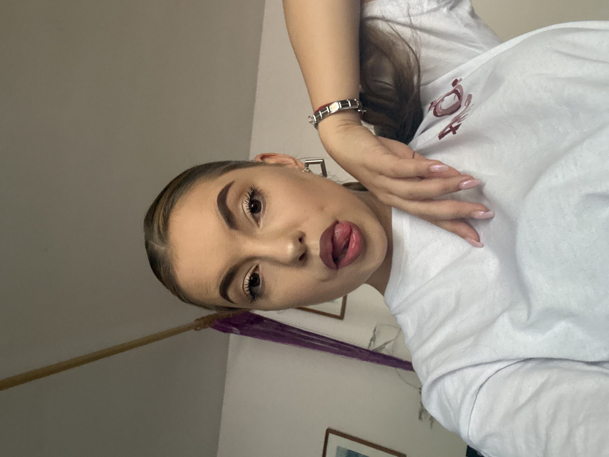 An indoor, medium shot shows a young woman with her tongue sticking out as she looks at the camera. She has her right arm resting on her chest, with her hand on her shirt. She is wearing a white t-shirt and a silver bracelet on her right wrist. The background shows a wall and a wooden pole. There is no indication of the location being in Sofia, Bulgaria.
