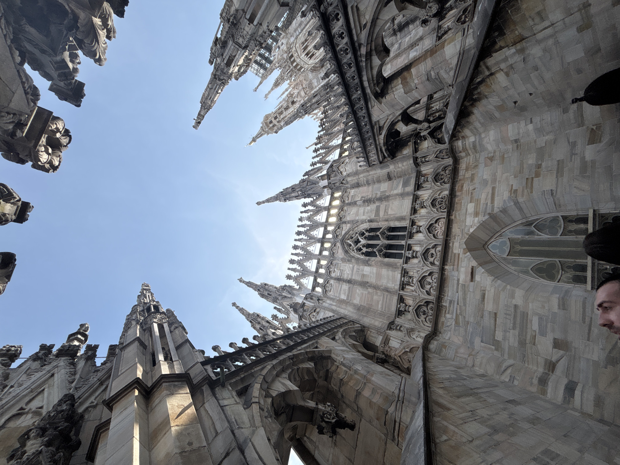 The image depicts the exterior architecture of the Duomo di Milano (Milan Cathedral) in Milan, Italy, viewed from a low angle looking upward. The stone structure is detailed with spires, pinnacles, arches, and ornate carvings. The sky is a pale blue, partially obscured by the cathedral's ornamentation. A portion of a man's face is visible in the bottom right corner of the frame. The architecture indicates the Gothic style of the cathedral.
