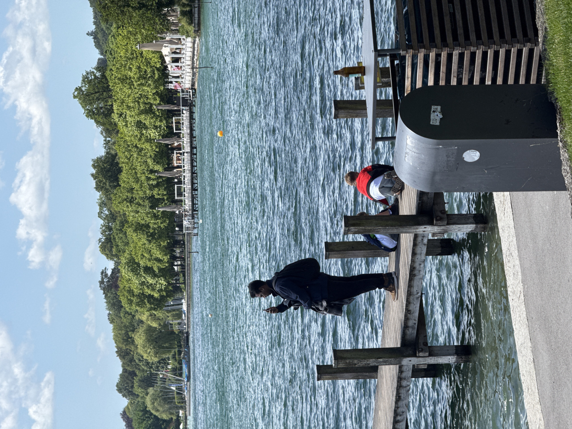 The image depicts a scene at Starnberger See, Germany, on a sunny day with scattered clouds. In the foreground, a dark grey, rounded trash receptacle stands to the right of a paved walkway. Adjacent to the trash can, a wooden structure made of horizontal planks is visible, potentially a seating area or a partition.
To the left of the trash can, a wooden pier extends into the blue, choppy water of the lake. On the pier, a man is standing, facing the water and holding a smartphone up to take a picture or video. He is wearing a dark jacket over a checkered shirt and dark pants, with a black backpack slung over his shoulders. He has a beard and dark hair. Further down the pier, two individuals are seated. One person is wearing a red and black jacket over a white shirt and grey pants, with their back to the camera, looking down at the pier. Another person, partially visible to the far right on the pier, is wearing a backpack. A brown glass bottle rests on a table on the pier to the right, near the seated individuals.
The background features a tree-lined shore with lush green foliage. Beyond the trees, a long white pier or boardwalk with umbrellas and seating can be seen, suggesting a public area or a restaurant. Sailboats are anchored in the distance on the left side of the lake. The water is a deep blue with whitecaps, indicating a breezy day.