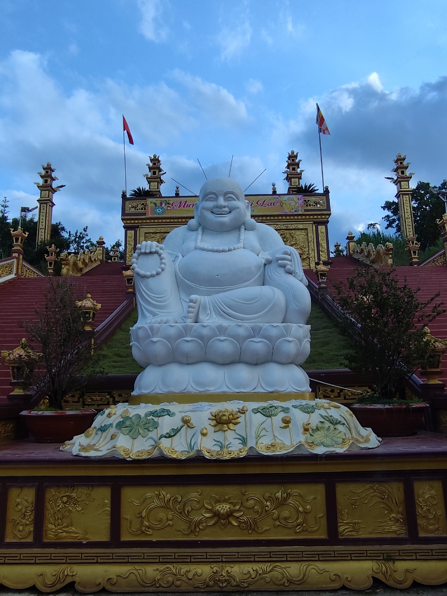 A large, white statue of the Laughing Buddha (Hotei or Budai) is the central focus of this image, set against the backdrop of a temple in Lạc Dương, Vietnam. The Buddha is depicted with a wide, jovial smile, a plump belly, and is seated on a multi-layered lotus pedestal. He holds a string of prayer beads in his right hand and a sack in his left.
The statue is positioned at the base of a grand religious complex featuring ascending red stairways flanked by green grass and traditional gold-colored lanterns. Above the stairs, a multi-tiered temple structure with intricate golden embellishments and decorative patterns is visible. Against a partly cloudy blue sky, two flags are raised on poles: one is a solid red flag, likely the Vietnamese national flag, and the other is a striped Buddhist flag.
On the golden structure directly behind the Buddha, partially visible Vietnamese text reads "Mừng Lạc." The Buddha statue rests on an ornate golden-colored base, decorated with detailed relief carvings of lotus flowers and other traditional motifs. Potted plants are placed around the statue and along the stairs. The scene is set outdoors during the daytime, likely late afternoon, conveying a sense of serenity and spiritual significance.