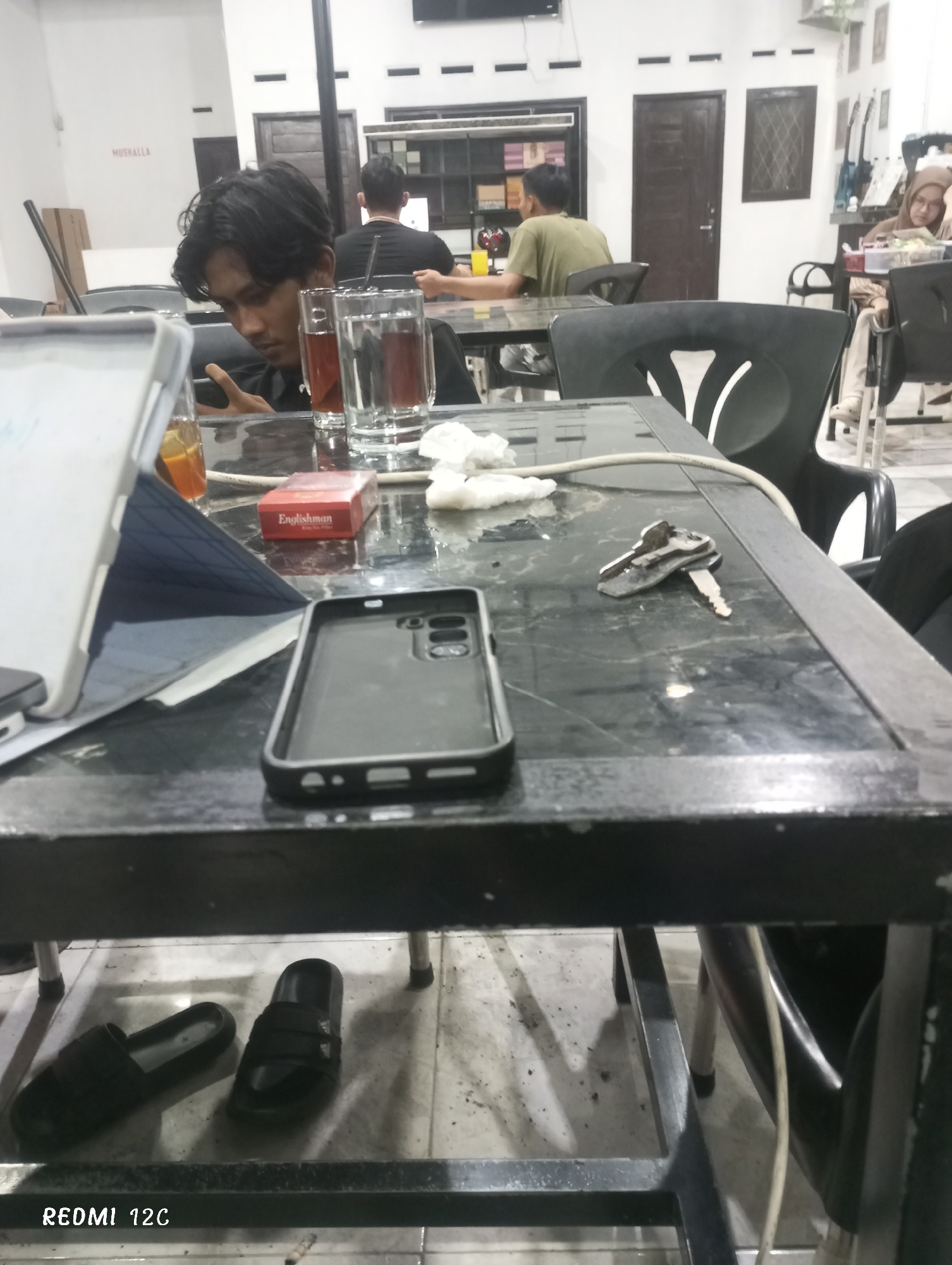 The scene is a dimly lit interior space, likely a cafe or restaurant in Lhokseumawe, Indonesia. A young man with dark hair is seated at a black marble-patterned table, absorbed in his mobile phone. On the table, along with a tablet, a phone case, keys, and crumpled tissue, is a pack of "Englishman" brand cigarettes, indicating that this is an area where smoking is permitted. Two glasses, one containing tea and the other water, are also on the table. The footwear – two black slides – are situated under the table.
In the background, there are other patrons seated at tables, including two men wearing dark clothing and another in an olive-colored shirt. Another table is occupied by a woman wearing a hijab. The furniture is black and generic, suggesting a budget-conscious establishment. Natural light enters the space, but it is limited, contributing to the dimness. The word "Mushalla" is printed on the wall. A door and window are visible in the background, lending context to the indoor setting.