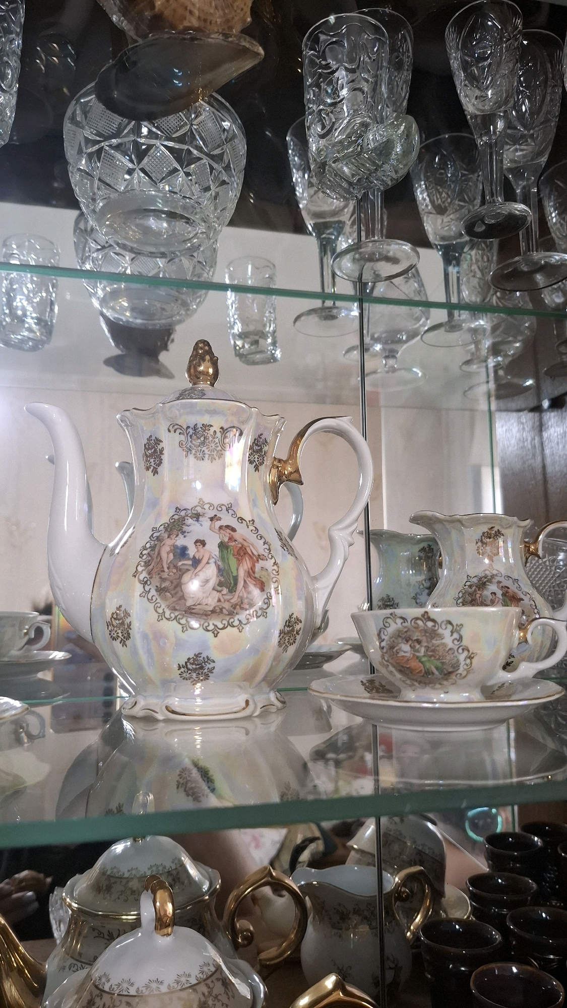 The image shows a collection of porcelain and glass items displayed on shelves within a glass-fronted cabinet. The primary subject is a white porcelain teapot adorned with intricate gold trim and a painted scene of figures in classical attire. To the right of the teapot are a porcelain creamer, a teacup and saucer, and a crystal decanter or pitcher. In the background, various crystal glasses and cups are visible, some with ornate patterns. The shelves are made of glass, and reflections of the objects and the surrounding environment are visible on their surfaces. The overall aesthetic suggests an antique or decorative display. The image does not provide any information or context related to the city of Shymkent, Kazakhstan, nor does it depict any people or actions.