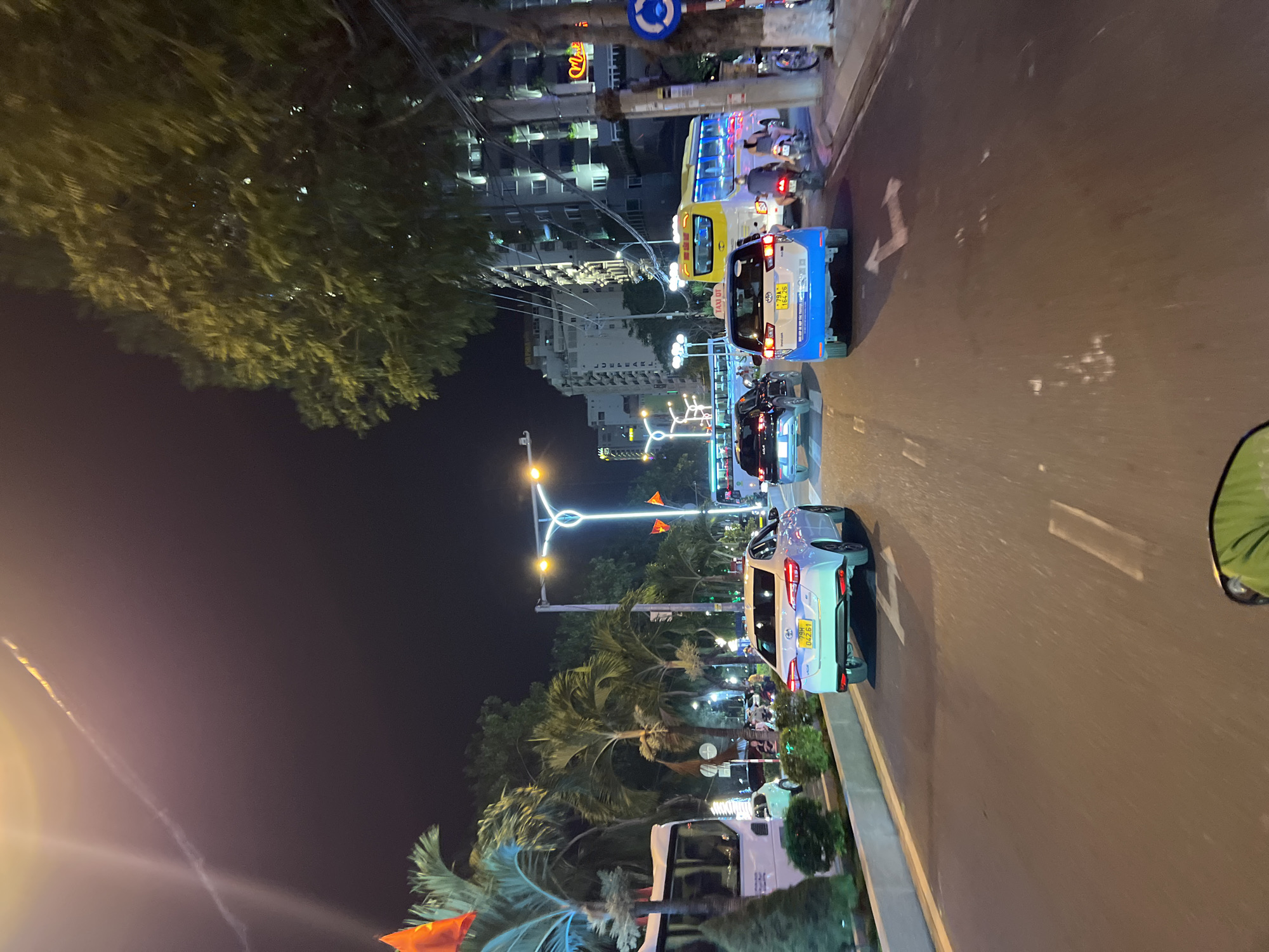 The image shows a nighttime street scene in Nha Trang, Vietnam. Traffic is moving along a multi-lane road with buildings visible in the background. Several cars and motorbikes are present. Streetlights illuminate the scene. There are trees and greenery lining the side of the road. There is a road sign visible. A Vietnamese flag is partially visible in the lower-left.