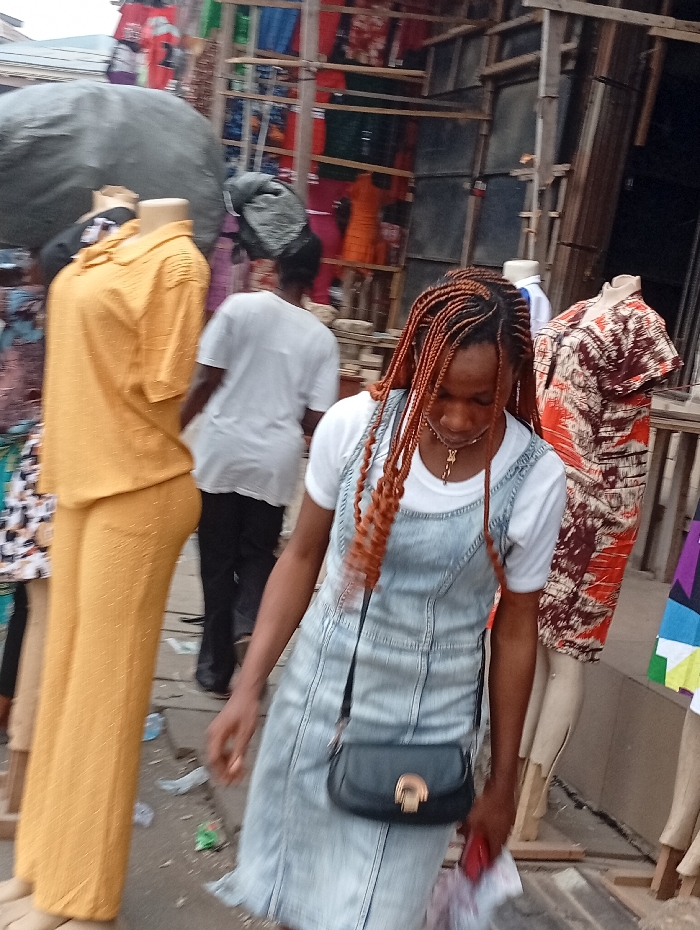 A woman with braided hair, wearing a light blue denim dress over a white t-shirt and a black purse, walks past mannequins dressed in clothes in an outdoor market setting. Colorful fabrics hang in the background. A person wearing a white shirt walks behind her. Other mannequins are visible with assorted dresses and clothing. The scene depicts a busy market atmosphere.
