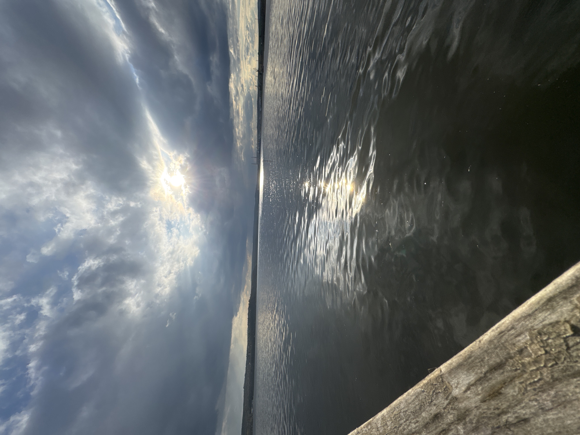 The image depicts a body of water, likely a lake or sea, under a cloudy sky with the sun partially obscured. The sun's reflection is visible on the water surface. A wooden structure, possibly a pier or dock, is partially visible in the foreground. The location is likely in or near Казашко, Bulgaria, suggested by the presence of a water body and potential coastal or lakeside environment in Bulgaria. There are no people visible in the scene. The focus is on the natural elements of water, sky, and sunlight, along with the man-made structure in the foreground.