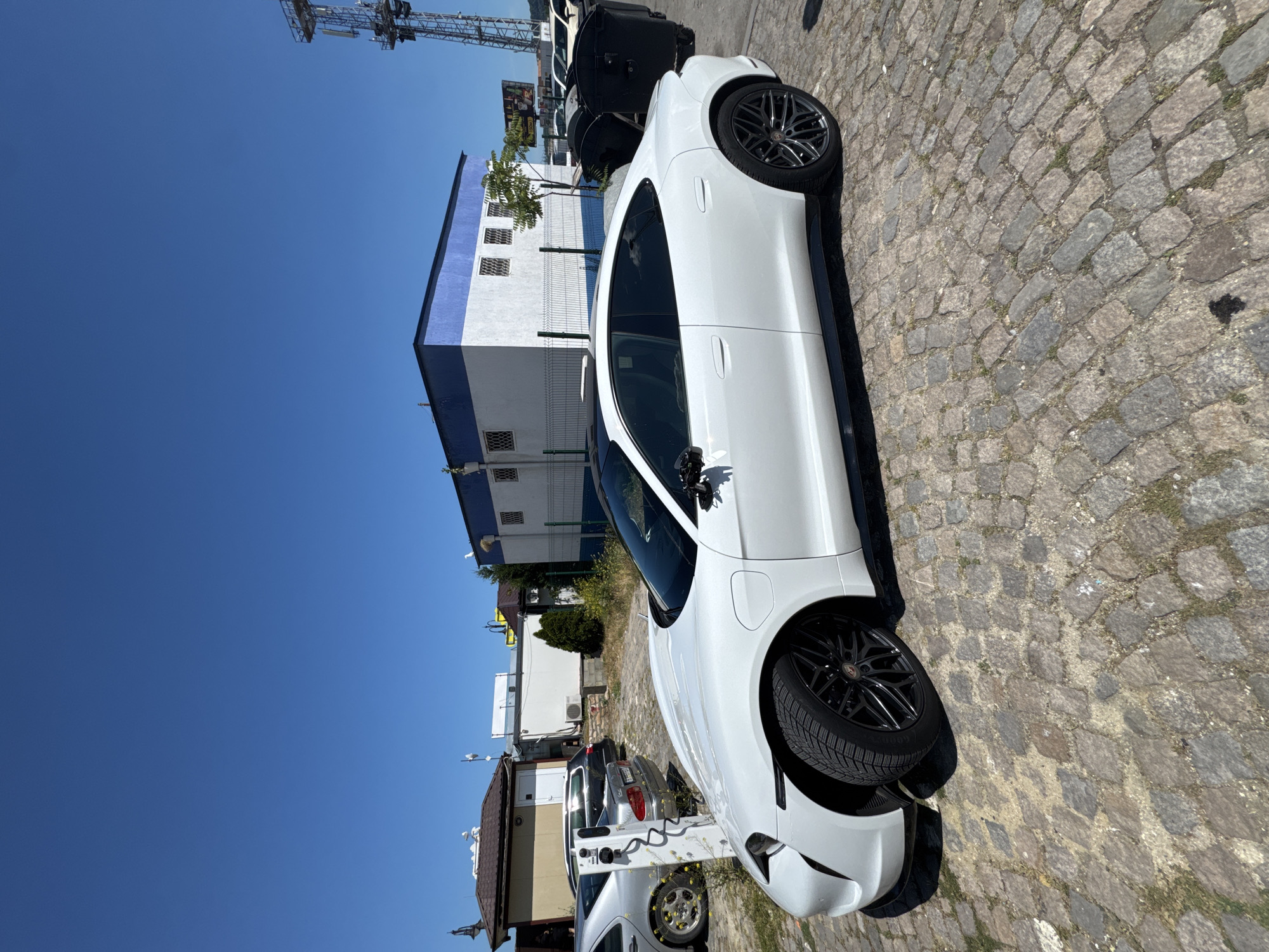 A white electric Porsche Taycan is parked on a cobblestone lot. It is being charged by a white charging station that is plugged into the car. In the background, there are several buildings and a cell tower. The sky is clear and blue. The location is in Varna, Bulgaria, near the port. There are no people visible in the image.