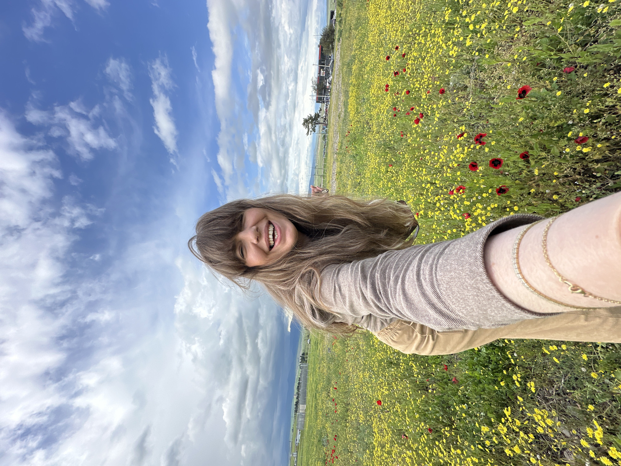 A woman is taking a selfie in a field of yellow and red flowers near Marneuli, Georgia. The sky is partly cloudy. A building, trees, and a fence are visible in the background. She is wearing a beige long-sleeved shirt and khaki pants. She is smiling. She has long brown hair with bangs. Two gold bracelets are visible on her left wrist.