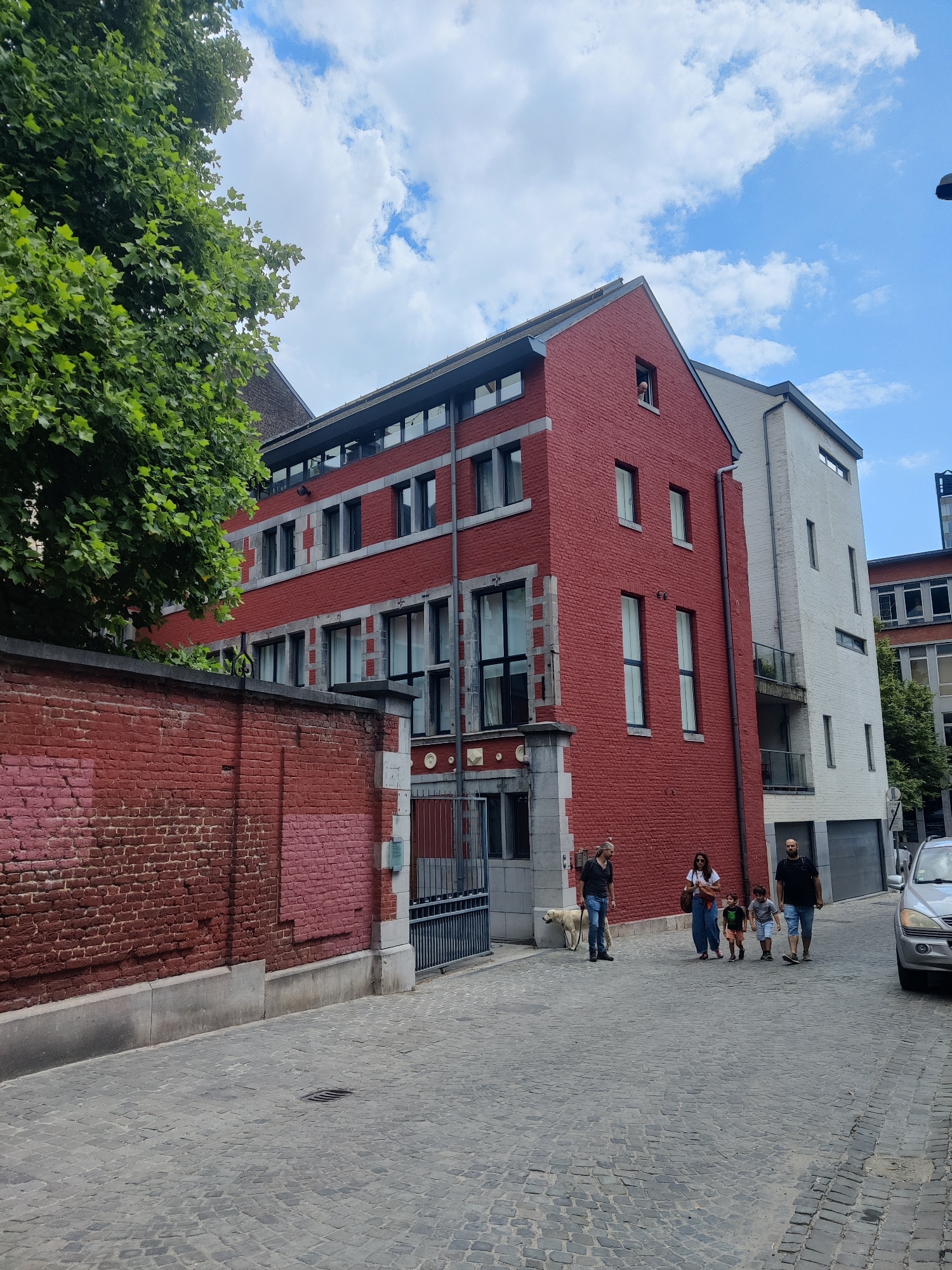 A family walks down a cobblestone street in Liège, Belgium, past a red brick building and a large tree. A man with a dog walks ahead of the family. The building has several windows and a red brick facade. A white building is next to the red brick building. The sky is cloudy and blue. A silver car is parked on the right side of the street.
