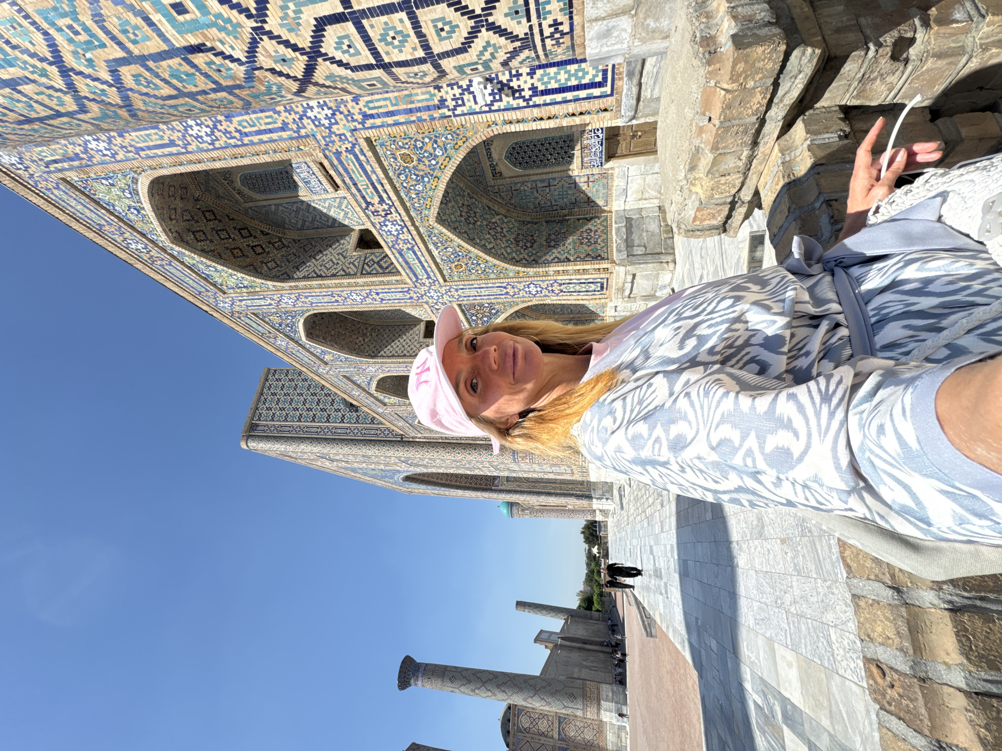 A woman in a pink cap and patterned robe poses for a photo in Samarkand City, Uzbekistan. She stands near a building adorned with intricate blue tilework and archways. In the background are pillars and pedestrians. The sky is clear and blue.
