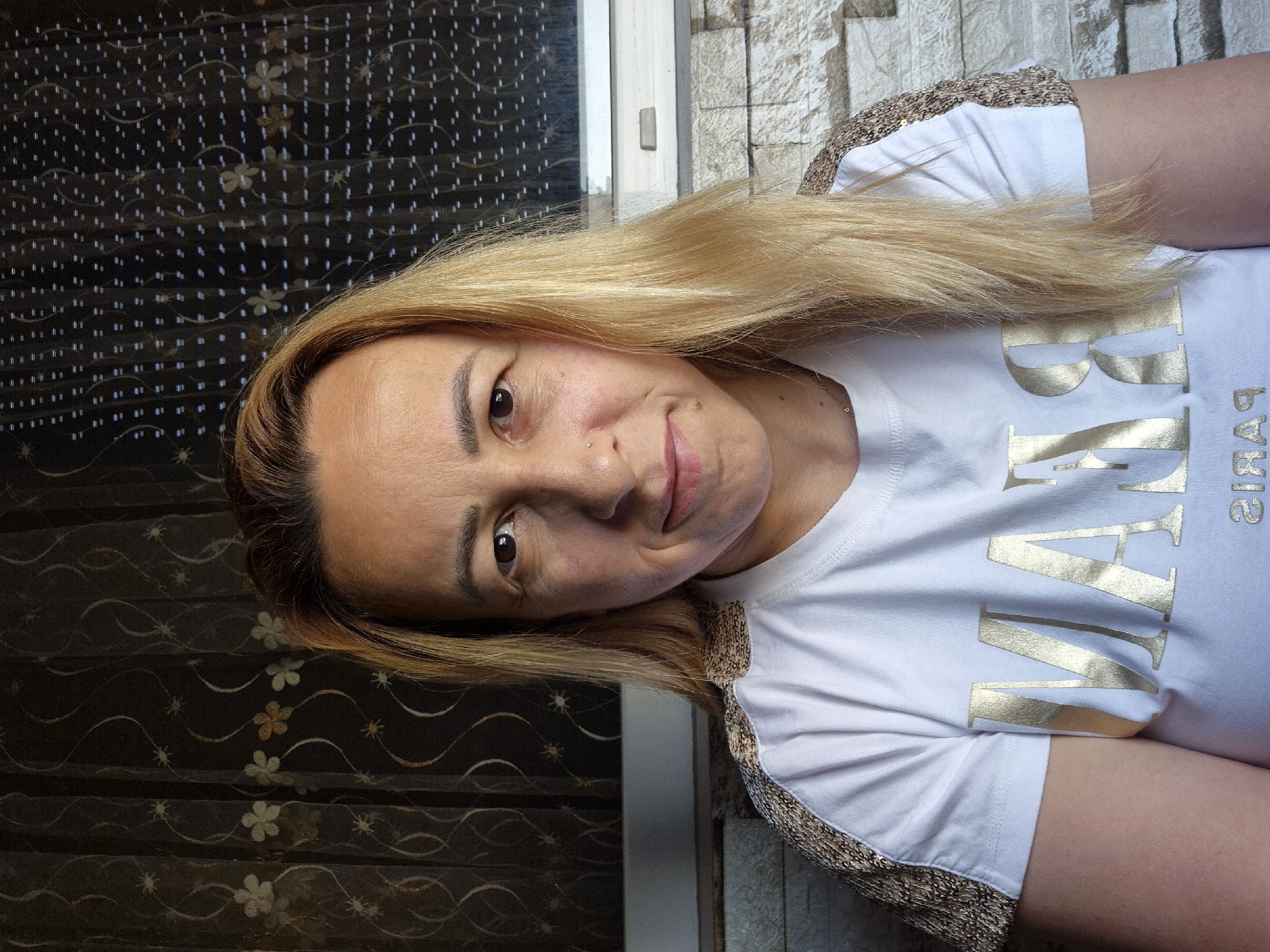 A medium shot shows a woman with blonde hair pulled back from her face, wearing a white t-shirt with gold lettering and gold sequined sleeves. She is looking directly at the camera and smiling slightly. The background consists of a dark curtain with floral patterns on the left and a window with blinds on the right. The lower portion of the image shows a stone wall. The scene is indoors, with soft lighting.