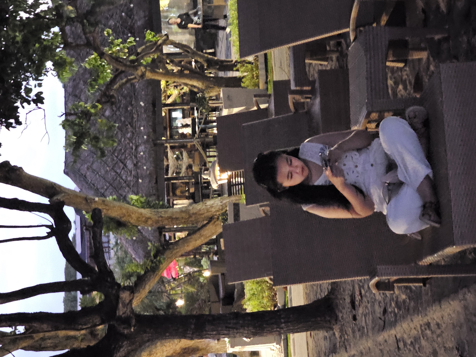 A young girl with long dark hair is sitting cross-legged on a lounge chair on a beach, looking down at her phone. She is wearing a white outfit. In the background, there are more lounge chairs, tables, trees, and a thatched-roof building. The scene appears to be at dusk. This setting is consistent with a beachside restaurant or resort in Denpasar, Indonesia.