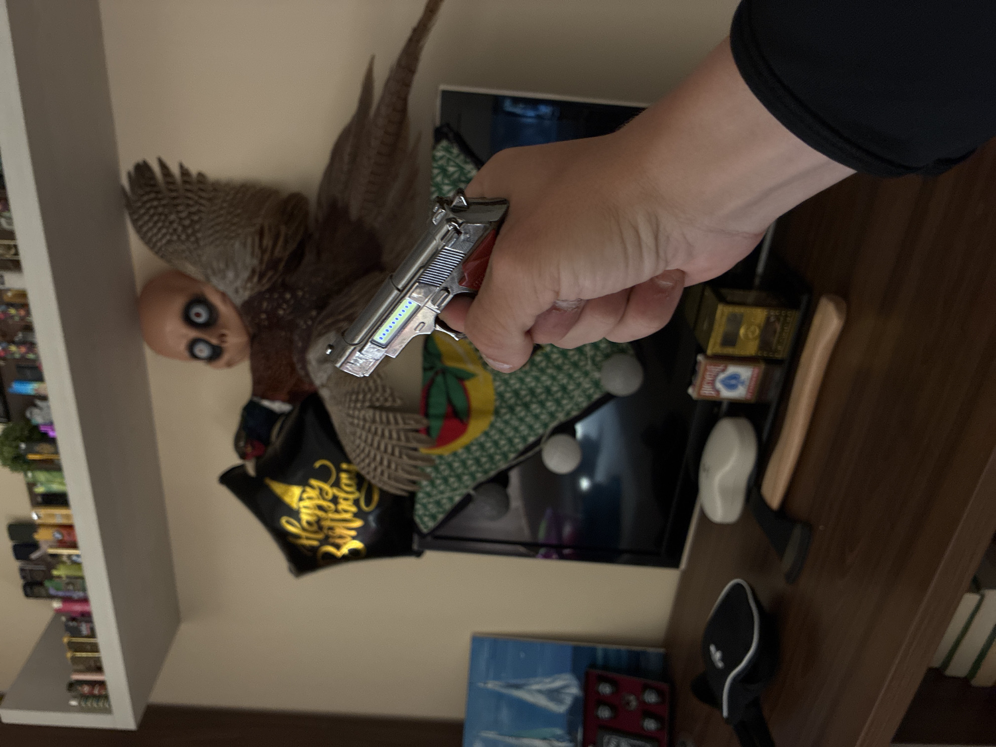 The image, originally rotated, displays an indoor scene dominated by a person's right hand holding a small, silver-colored pistol in the foreground. The hand extends from the right of the frame, revealing a black sleeve on the wrist. The pistol features red accents on its grip and a row of illuminated blue lights along its side, suggesting it is a toy, a replica, or a functional item like a lighter.
Behind the hand and pistol is a unique doll-like figure. It has a bald, pale head with prominent, wide-open black and white eyes. This head is attached to a body adorned with large, brown feathered wings, giving it the appearance of a peculiar, possibly unsettling, angel-like creature or a taxidermied bird with a doll's head.
The background shows a cluttered shelf or cabinet. A television screen is partially visible behind the doll, along with a green-patterned cushion. On the wooden surface, several items are arranged, including a small axe with a wooden handle, a black baseball cap, and a box of "Bicycle" brand playing cards. To the far left, a white shelving unit is filled with numerous small, colorful objects. A black balloon with the text "Happy Birthday" in gold script is also visible near the feathered figure. The scene is illuminated by artificial light, indicating it is indoors. No specific cues within the image indicate its geographical location.