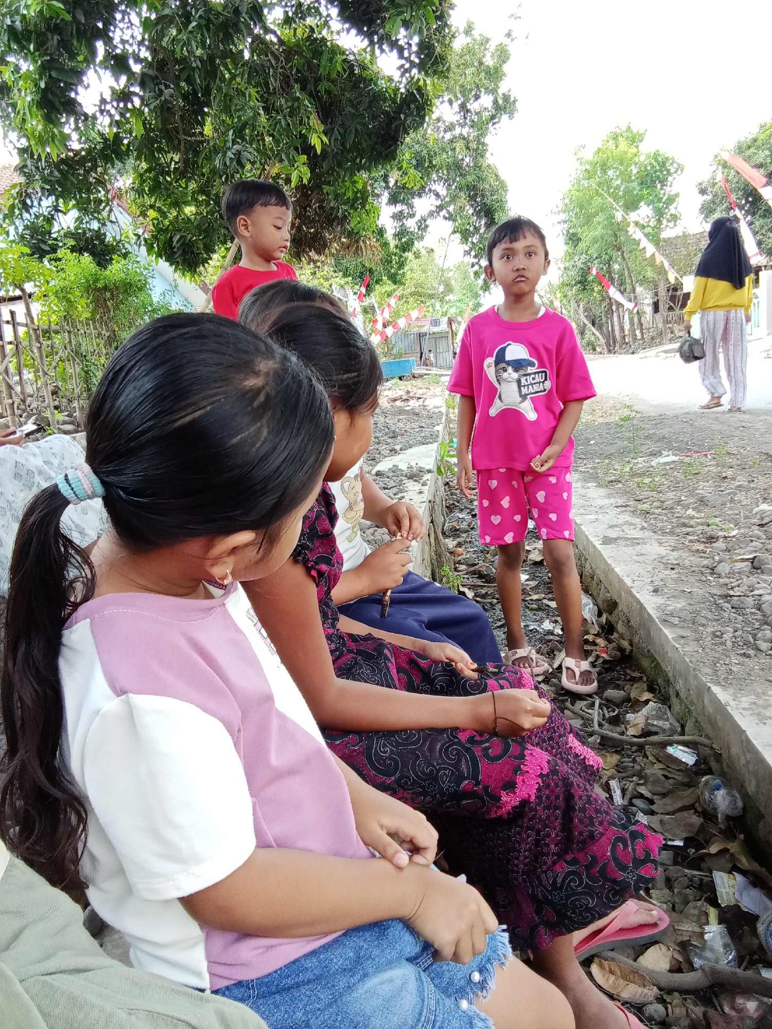 The image depicts an outdoor scene with several individuals, likely in Trebungan, Indonesia, given the context. In the foreground, two young girls are seated, facing away from the camera. The girl on the left has long, dark hair tied in a ponytail with a blue and light pink elastic band. She is wearing a t-shirt with pink and white color-blocking and blue denim shorts. The girl next to her is wearing a dark purple dress with a black and pink floral pattern. Her hands are visible, engaged in some activity.
Standing in the middle ground is a young boy in a bright pink t-shirt with a graphic of a cat wearing a baseball cap and the text "KICAU MANIA" (Bird Lovers). He is also wearing pink shorts with a heart pattern and light pink sandals. His expression is directed towards the camera. Further in the background, another young boy in a red t-shirt is partially visible, peeking over the seated girls. To the right, a woman wearing a yellow hooded jacket and a black hijab is walking away from the camera, dressed in striped pants.
The environment appears to be a street or pathway with a concrete edge. The ground is covered with dirt, leaves, and some scattered debris, including plastic bottles and wrappers. Lush green trees and foliage are abundant, suggesting a rural or semi-urban setting. Decorative red and white flags or banners are visible on poles in the background, possibly indicating a local celebration or event. The overall lighting suggests it is daytime.