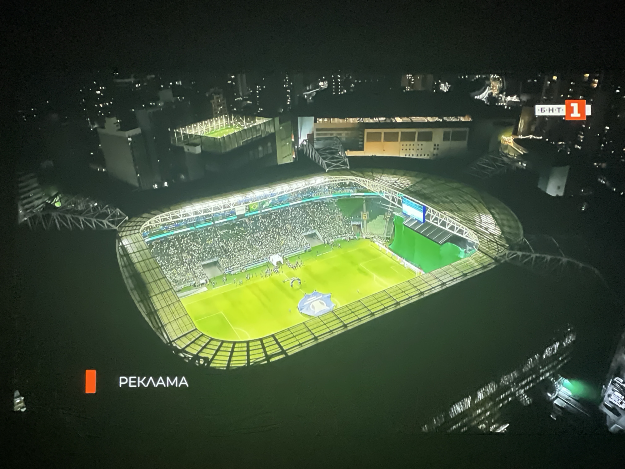 The image shows an aerial view of a brightly lit football stadium at night, likely in a city. The stadium itself is a large, oval-shaped structure with a green field in the center and tiered seating filled with spectators. The stadium is surrounded by other buildings and lights, indicating it is located within an urban area. A logo with a red square and the number "1" is visible in the upper right corner of the image, along with Cyrillic text that appears to read "BHT". In the lower left corner, an orange rectangle is displayed next to the white text "РЕКЛАМА," which translates to "ADVERTISEMENT" in English. The overall scene suggests a broadcast of a sporting event, possibly with an advertisement break. There is no information in the image that specifically places it in Ruse, Bulgaria.