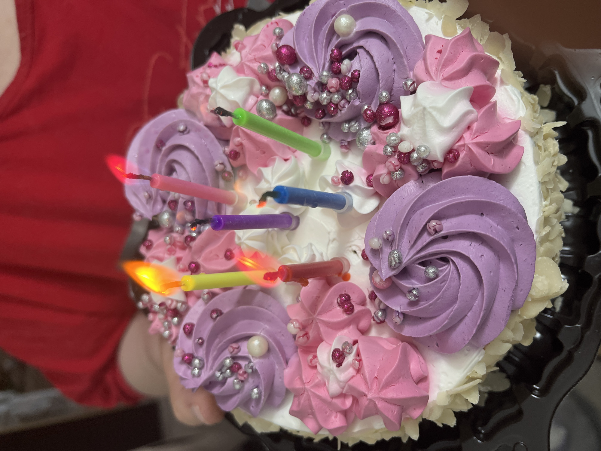 The image shows a decorated birthday cake with lit candles. The cake has white icing and is decorated with swirls of purple, pink, and white frosting. Silver and pink edible decorations are scattered across the top. Seven candles of different colors (green, pink, blue, purple, yellow, red) are inserted into the cake, with some burning. A person wearing a red shirt is visible on the left side of the image, holding the cake in its black plastic container. The location in the city of Chaikovsky, Russia is not discernible from the image.