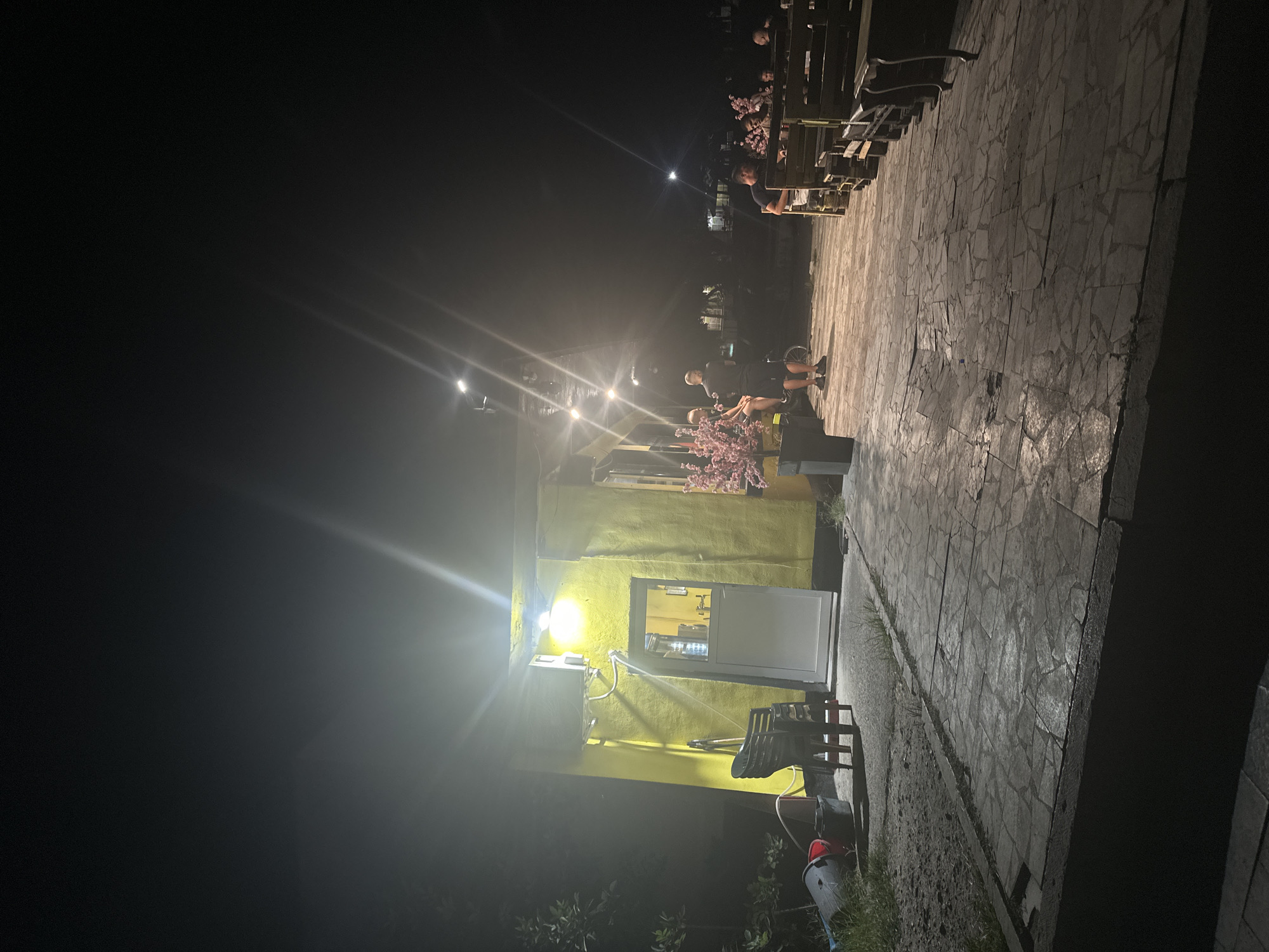 This is a night-time exterior photograph of a small, yellow, single-story building with a white door and window, situated in the city of Galiche, Bulgaria. The building appears to be a shop or cafe. A bright, overhead light illuminates the front of the building, casting strong glare and lens flare. In front of the building is a paved outdoor seating area with stone tiles. To the left of the building, on the grass, are several large bins and stacked chairs.
On the paved area in front of the building, there are several people. Two individuals are standing near the entrance, one of whom is looking at a bicycle. Further to the right, a group of people are seated at tables made of wooden pallets. One person is seated at a table, appearing to be dining, while other individuals are seated at tables further back, some engaged in conversation. Two decorative pink floral displays in black planters are positioned near the seating area. The overall scene suggests an outdoor gathering or dining space operating at night.