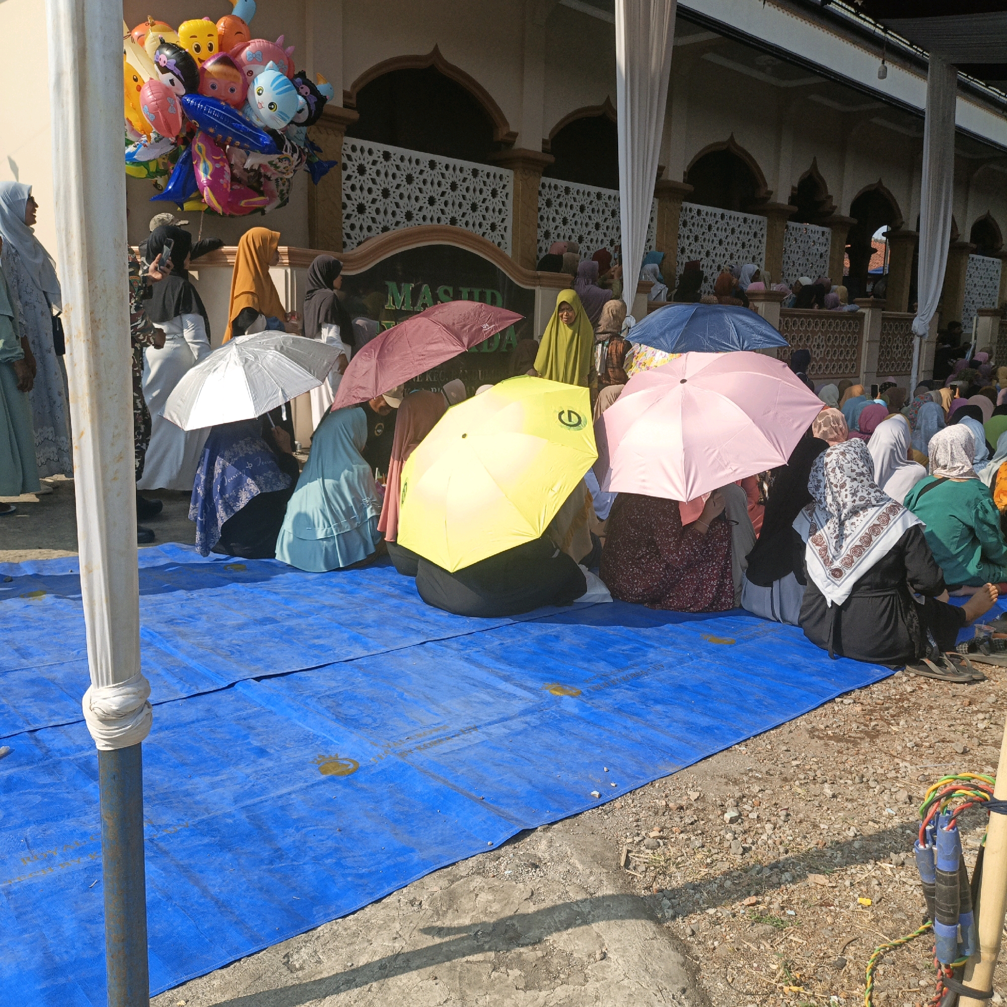In Kalitorong, Indonesia, a group of people, predominantly women dressed in hijabs, are gathered outdoors on a bright, sunny day, seated on a large blue tarp spread over a gravel and concrete surface. Many are using umbrellas for shade, with colors including pink, yellow, and maroon. A cluster of brightly colored balloons, featuring cartoon characters and animal designs, floats on the left side of the frame, attached to a white pole. In the background, a building with arched windows and ornate white latticework is visible, with white drapery hanging down. The scene appears to be a public gathering or event, possibly related to a religious occasion given the setting and attire.