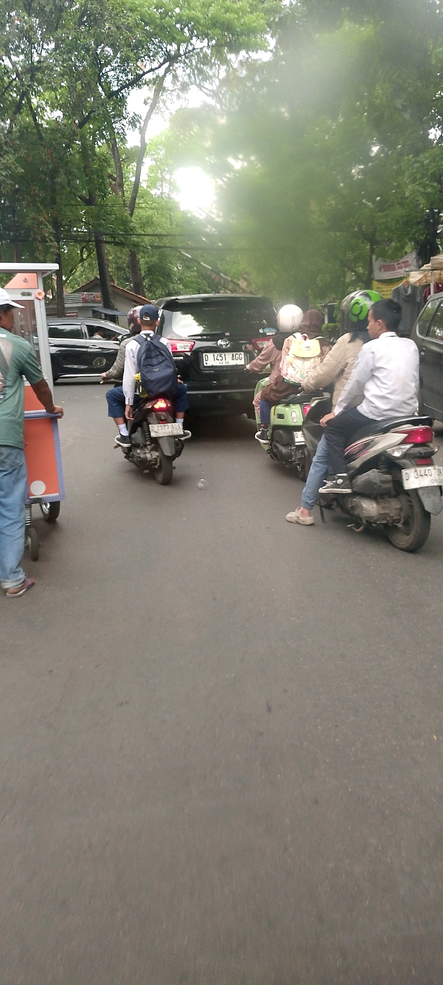 In an outdoor urban scene, likely Bandung City, Indonesia, a traffic jam is occurring on a paved road lined with trees. The sunlight is diffused and hazy, suggesting overcast or humid conditions. Several vehicles are present: a black SUV, at least two motorcycles, and a mobile food cart.
On the food cart, a person wearing a green t-shirt and blue jeans stands with their back to the camera, facing left.
Two individuals are on a motorcycle in the center of the frame. The person in front is wearing a blue baseball cap and a white shirt, with a blue backpack on their back. They are facing away from the camera. The person behind them is wearing a blue shirt and is also facing away.
To the right, another motorcycle has two individuals. The person in front wears a light brown hijab and a brown top, with a light yellow backpack on their back, facing away. The person behind them wears a light brown jacket and jeans, also facing away.
A third motorcycle, on the far right, has one person wearing a white shirt and dark pants, facing away from the camera, with their left leg extended forward.
The black SUV has a license plate that reads "D 1451 AGG". The motorcycle in the center has a license plate that reads "D 2573 BDA". The motorcycle on the right has a license plate that reads "D 3440 OH".
The environment appears to be a busy street with trees providing shade, and the road surface shows some wear and tear. The overall impression is of a typical day in a bustling Indonesian city.