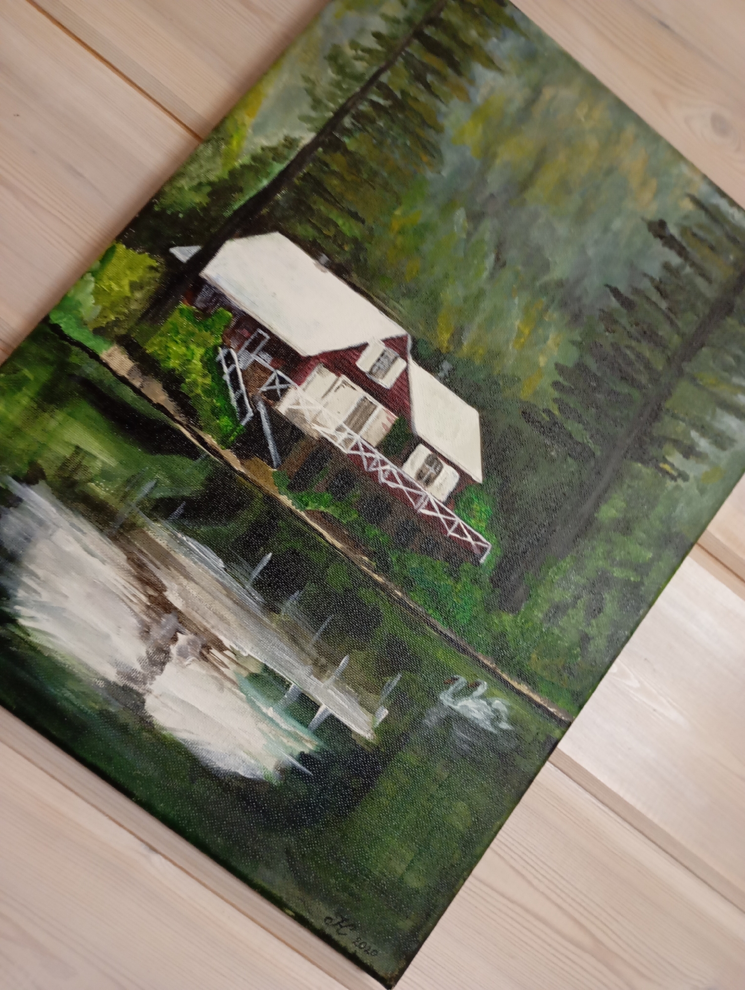 A painting depicts a red and white house situated amongst trees, with a body of water in the foreground. The house has a white roof and a red facade with white trim around the windows and a white railing on a small porch. The water reflects the trees and the house, and two white swans are visible in the water. The painting is signed "K. 2010" in the bottom right corner. The painting is displayed on a light-colored wooden surface, likely a wall or table, with visible wood grain. The painting is oriented diagonally on the surface. No specific location in the city of Malyi Bukor, Russia, can be determined from the image.