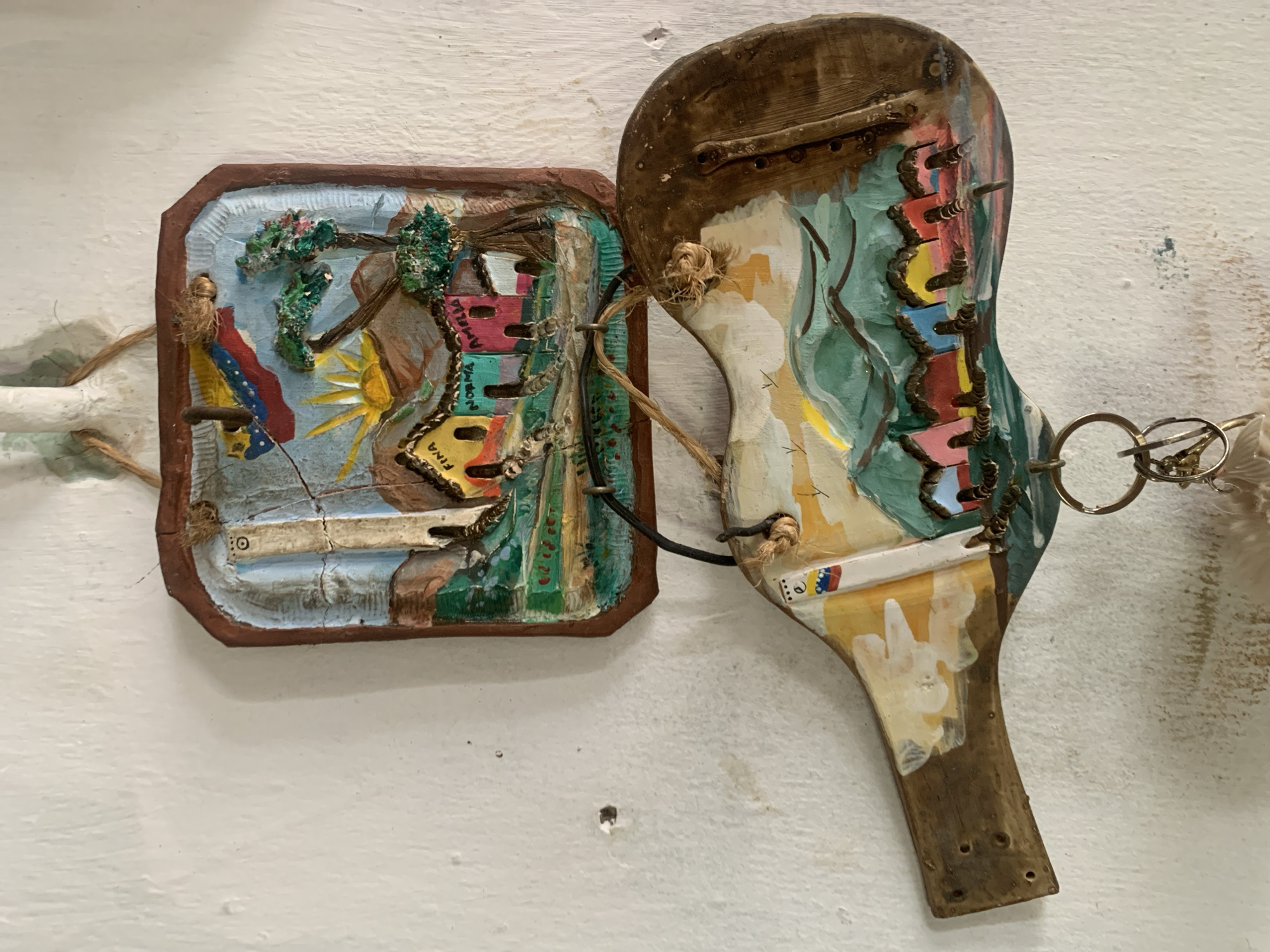 Two pieces of artwork are mounted on a white wall.
The first, on the left, is a rectangular, framed relief sculpture depicting a landscape. It features elements such as trees, a sun, and possibly houses, with the Venezuelan flag incorporated. The frame is brown.
The second, on the right, is an oddly shaped piece, perhaps resembling a stylized paddle or racket. It is also painted and includes metallic components, such as rings. It appears to incorporate wood and metal elements in its construction.
The artwork is connected by what appears to be twine or cord.
The overall aesthetic suggests folk art or outsider art, possibly related to the city of Santa Ana, Venezuela, based on the inclusion of the flag.