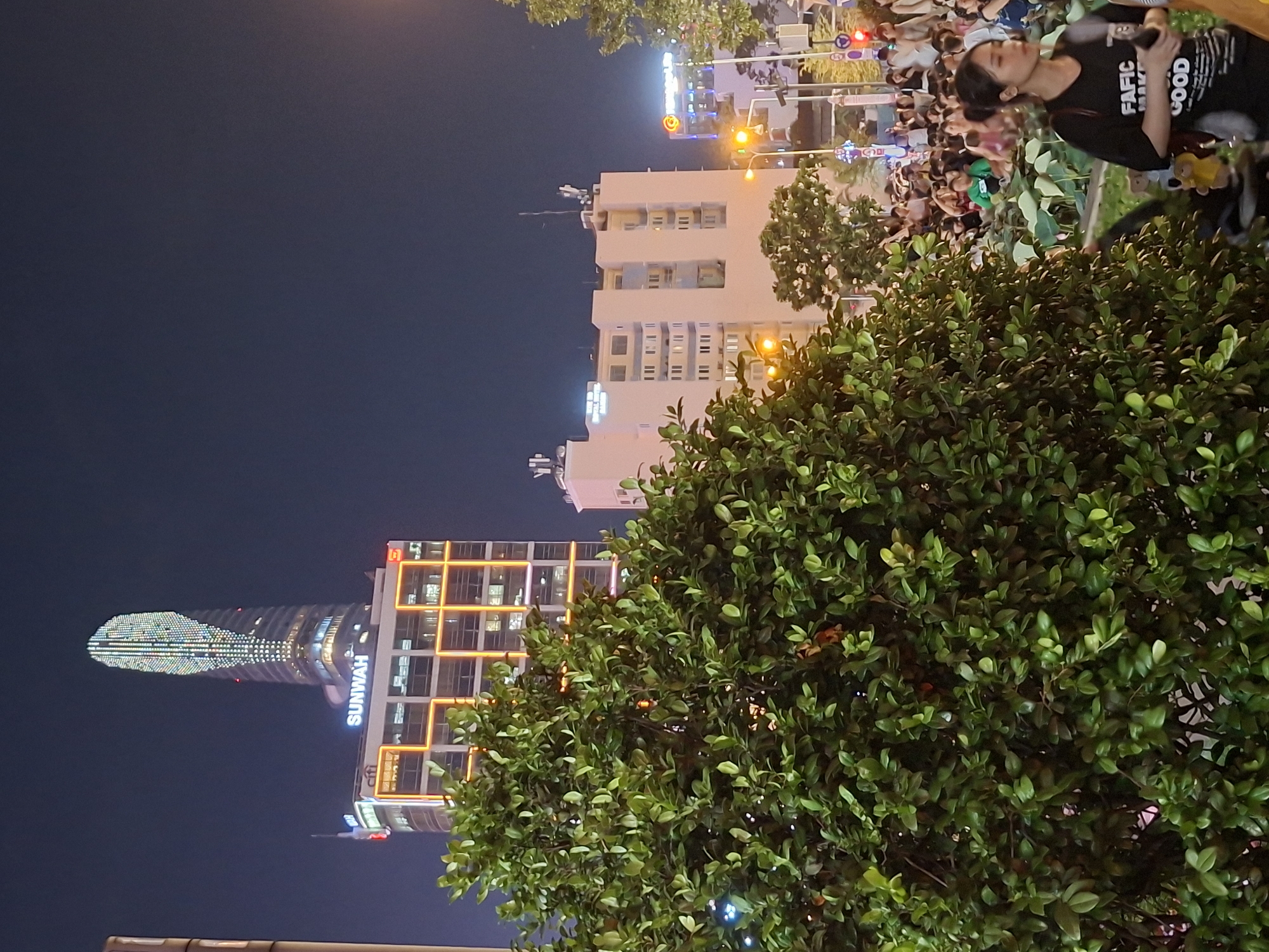 The image depicts a nighttime street scene in Thủ Đức City, Vietnam. In the background, the distinctive illuminated upper section of the Landmark 81 skyscraper is visible, with the "SUNWAH" sign illuminated on a lower building. Several other buildings, including a light-colored building with multiple windows and antennas, are also present. The foreground is dominated by a large, lush green tree with dense foliage. A crowd of people is gathered in the mid-ground, some appearing to be walking or standing in an open area, possibly a park or plaza. A traffic light with red and yellow indicators is visible on the right side of the frame. A young woman wearing a black t-shirt with white text, including "FIFIC GOOD," is in the right foreground, looking to her left and holding a dark object. The overall atmosphere suggests a bustling urban environment with illuminated architecture and a public gathering.