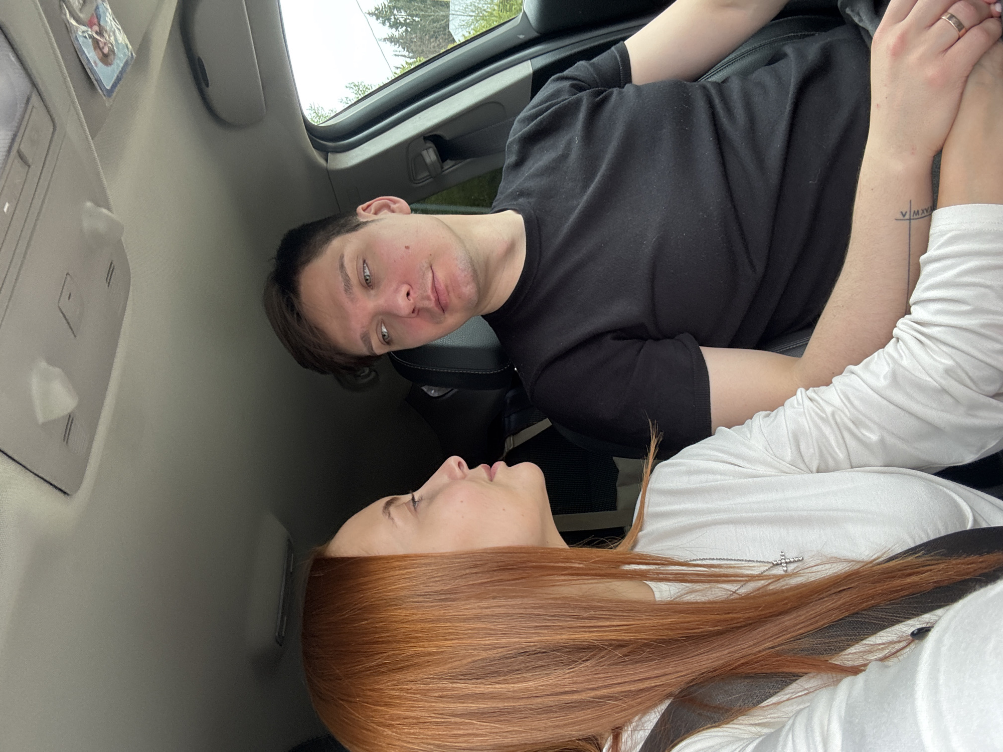 A man and woman are in a car. The man has short brown hair and is wearing a black t-shirt. The woman has long red hair and is wearing a white shirt. They are holding hands. The man has a tattoo on his arm. The location is Chastyye, Russia.