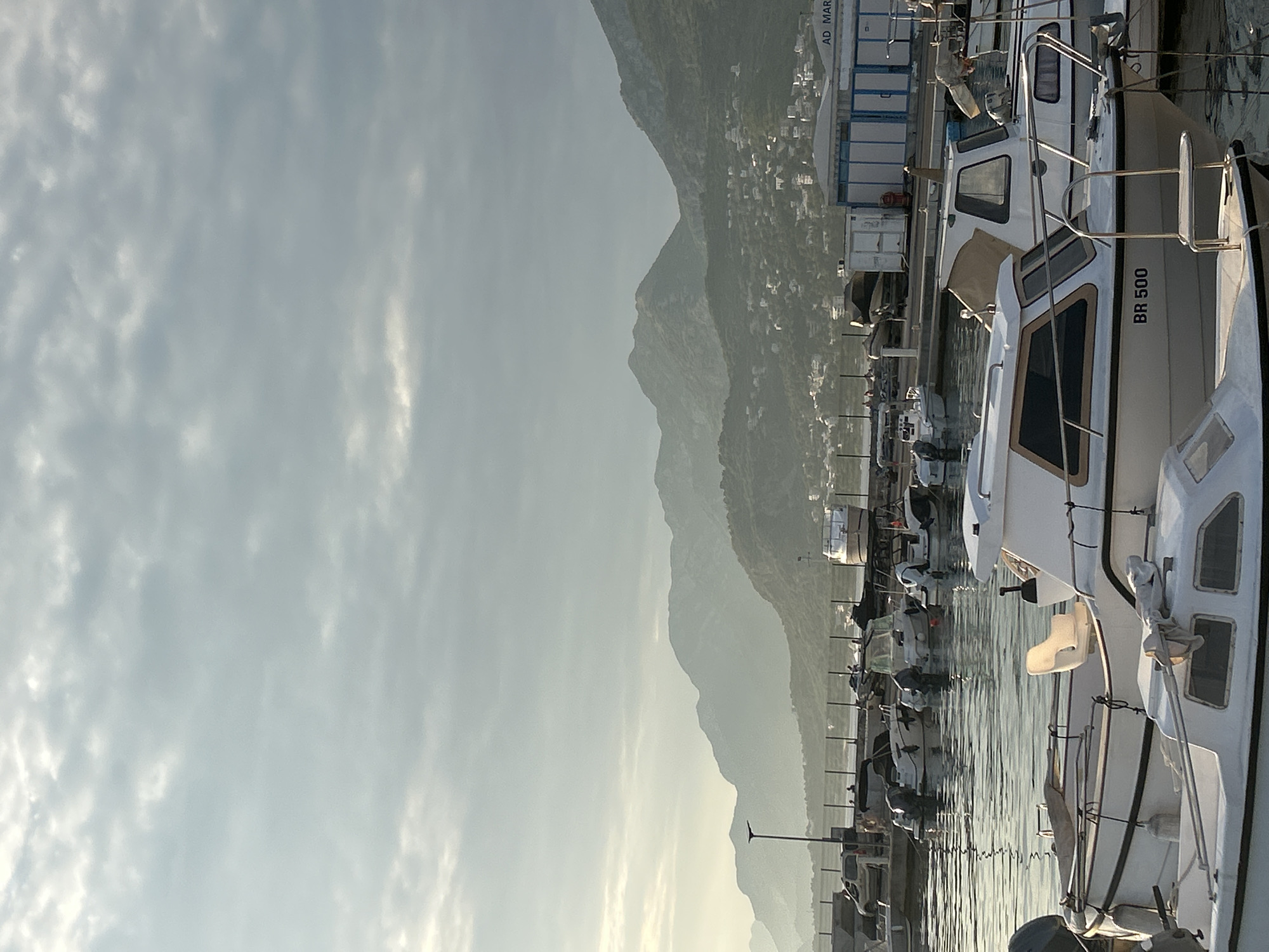 This image depicts a serene harbor scene in the city of Bar, Montenegro, likely at sunrise or sunset, given the soft, diffused light. Several white motorboats of varying sizes are docked in the calm, reflective water. In the foreground, two boats are prominently featured; one has the registration "BR 500" visible on its hull. Further back, a line of boats is moored along a dock, with a few more vessels visible in the distance. Behind the harbor, a row of buildings, possibly part of the port infrastructure, stands against a backdrop of rolling hills and distant, hazy mountains. The sky above is overcast with soft clouds, and the overall atmosphere is tranquil and quiet.