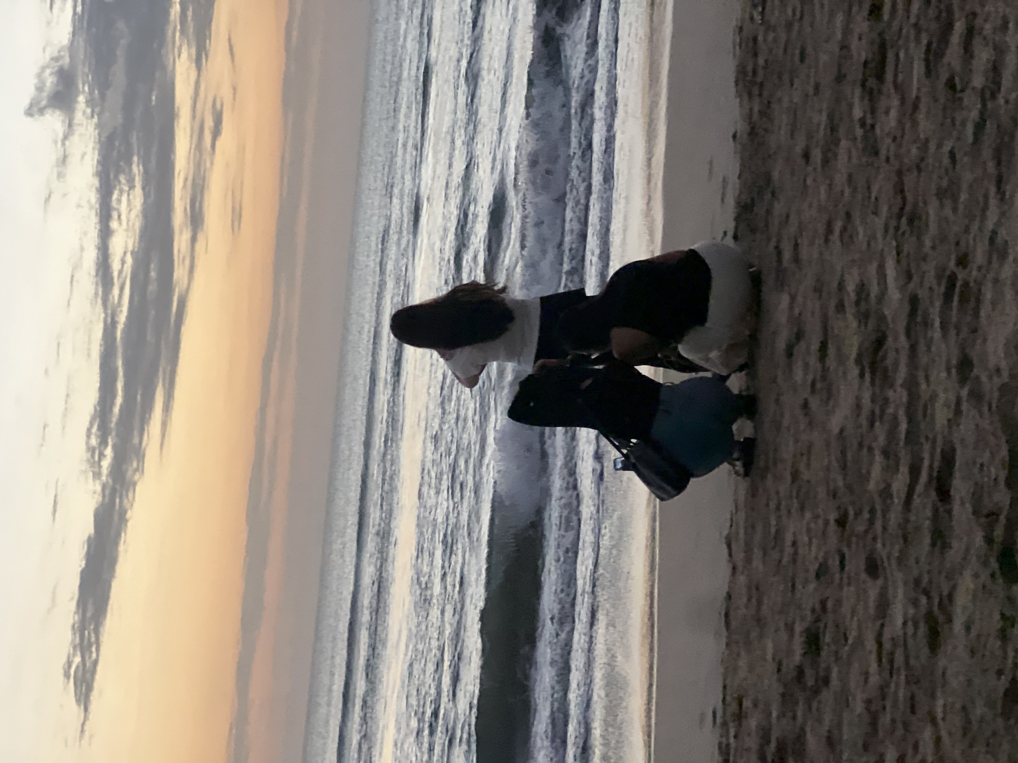 The image captures a serene sunset scene at Kuta Beach in Bali, Indonesia. Three individuals are gathered on the sandy shore, facing the ocean. One person stands slightly apart, their back to the camera, wearing a white top and dark skirt, seemingly observing the sunset. The other two individuals are crouched closer to the water, their backs also to the viewer. One of them is wearing a black top and light-colored bottoms, while the other is in a black top and blue jeans, with a dark bag at their side. The gentle waves of the ocean are visible, with a soft sunset sky painted in hues of orange, pink, and pale yellow. The atmosphere is tranquil and contemplative, typical of a peaceful evening on this popular Balinese beach.