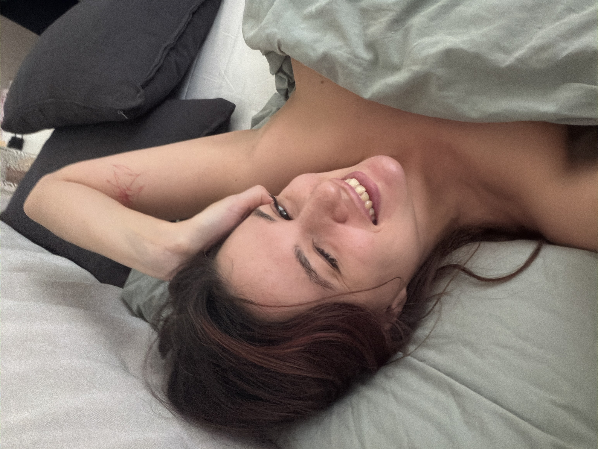 A woman is lying in bed, partially covered by a gray duvet. She is on her back, angled towards the camera. Her head is tilted, and she is smiling. Her hair is dark brown with red highlights. Her left arm is raised, with her hand near her head. The woman has markings or wounds on her left arm. There are two dark gray pillows behind her, resting on a white surface. The location is presumed to be an interior space within Belgrade, Serbia.