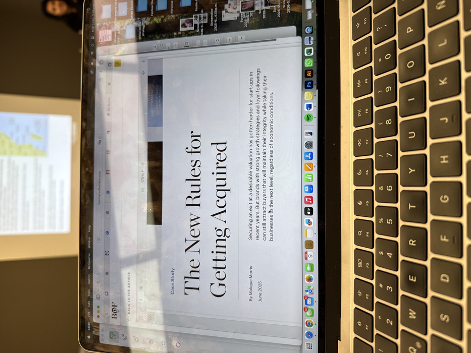 A laptop screen displays an article titled "The New Rules for Getting Acquired," by Malique Morris, published in June 2025. The article discusses the increasing difficulty of securing a desirable exit valuation for start-ups. It suggests that brands with strong growth strategies and loyal followings can still attract buyers who will maintain their integrity while advancing their businesses, regardless of economic conditions. The laptop's keyboard is visible on the right side of the image, and application icons are displayed on the dock at the bottom of the screen.