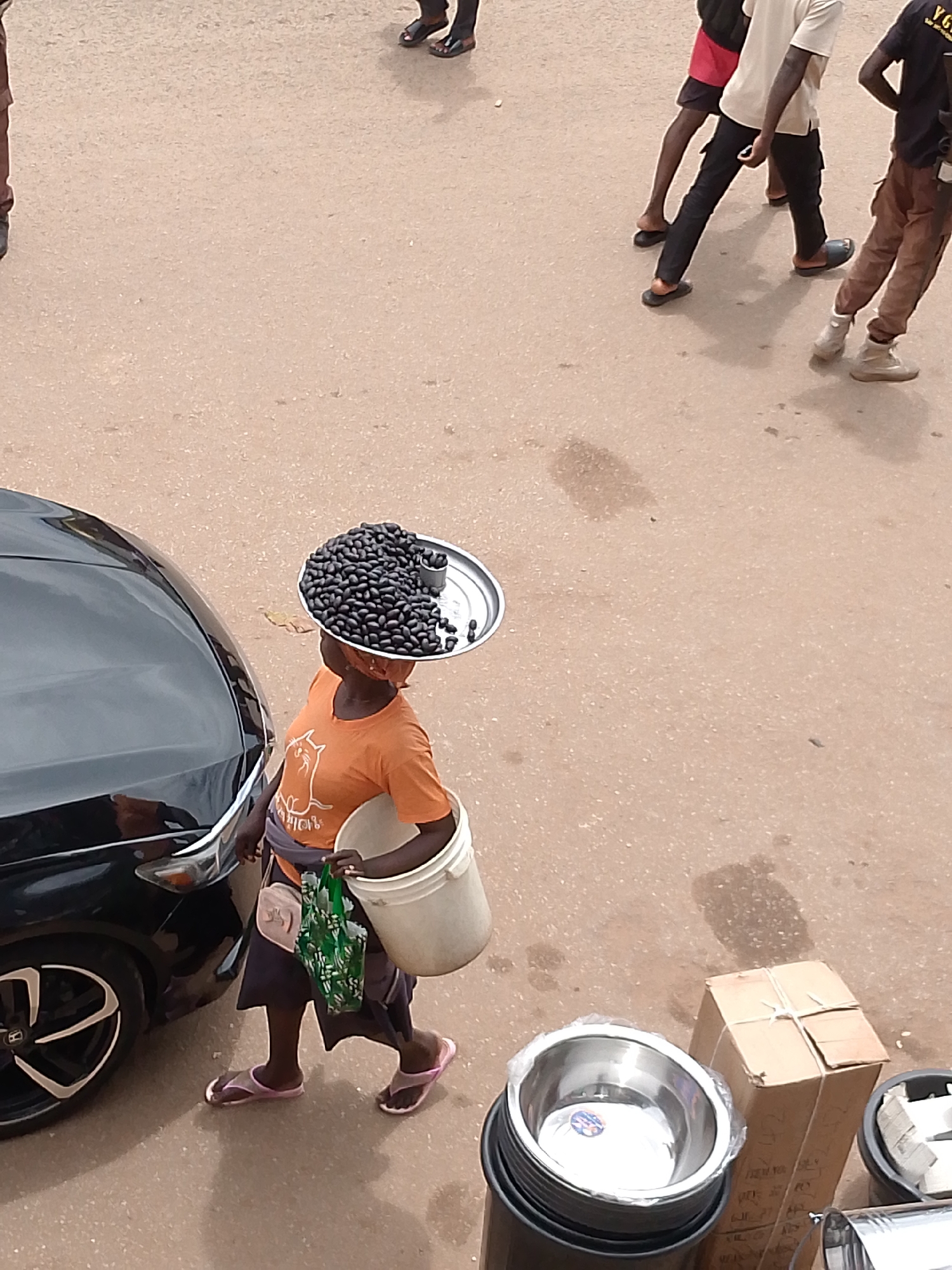 A high-angle photograph captures a street scene in Bukuru, Nigeria, during daylight hours. The ground is a light brown, textured surface, consistent with asphalt or packed earth.
In the mid-ground, a female individual walks from left to right, facing partially away from the camera. She wears an orange t-shirt featuring a white cat graphic, a dark skirt, and pink flip-flops. A reddish fabric is wrapped around her head. She precisely balances a circular silver tray on her head, which is piled high with numerous small, dark, oblong objects, resembling seeds, and contains a smaller silver measuring cup. In her left hand, she carries a white plastic bucket, and in her right hand, a green plastic bag. A small pink pouch is attached to her hip area.
To the left of the woman, the front quarter panel and wheel of a black sedan, bearing a Honda emblem on the wheel, are visible, appearing stationary.
In the upper right background, three male figures walk from right to left. One figure wears a light-colored t-shirt, dark trousers, and dark flip-flops. Another wears a dark t-shirt with visible yellow "YG" lettering, light brown cargo pants, and light-colored high-top footwear. A third figure, partially visible, wears a red garment and dark legwear with dark flip-flops. Shadows from these individuals are cast distinctly to their right.
In the lower right foreground, a brown cardboard box, secured with white string, rests on the ground. Adjacent to it are stacked black plastic buckets containing multiple metallic, likely stainless steel, bowls. One top bowl is covered with clear plastic and displays a blue circular sticker. A part of another metallic container is also visible on the ground.