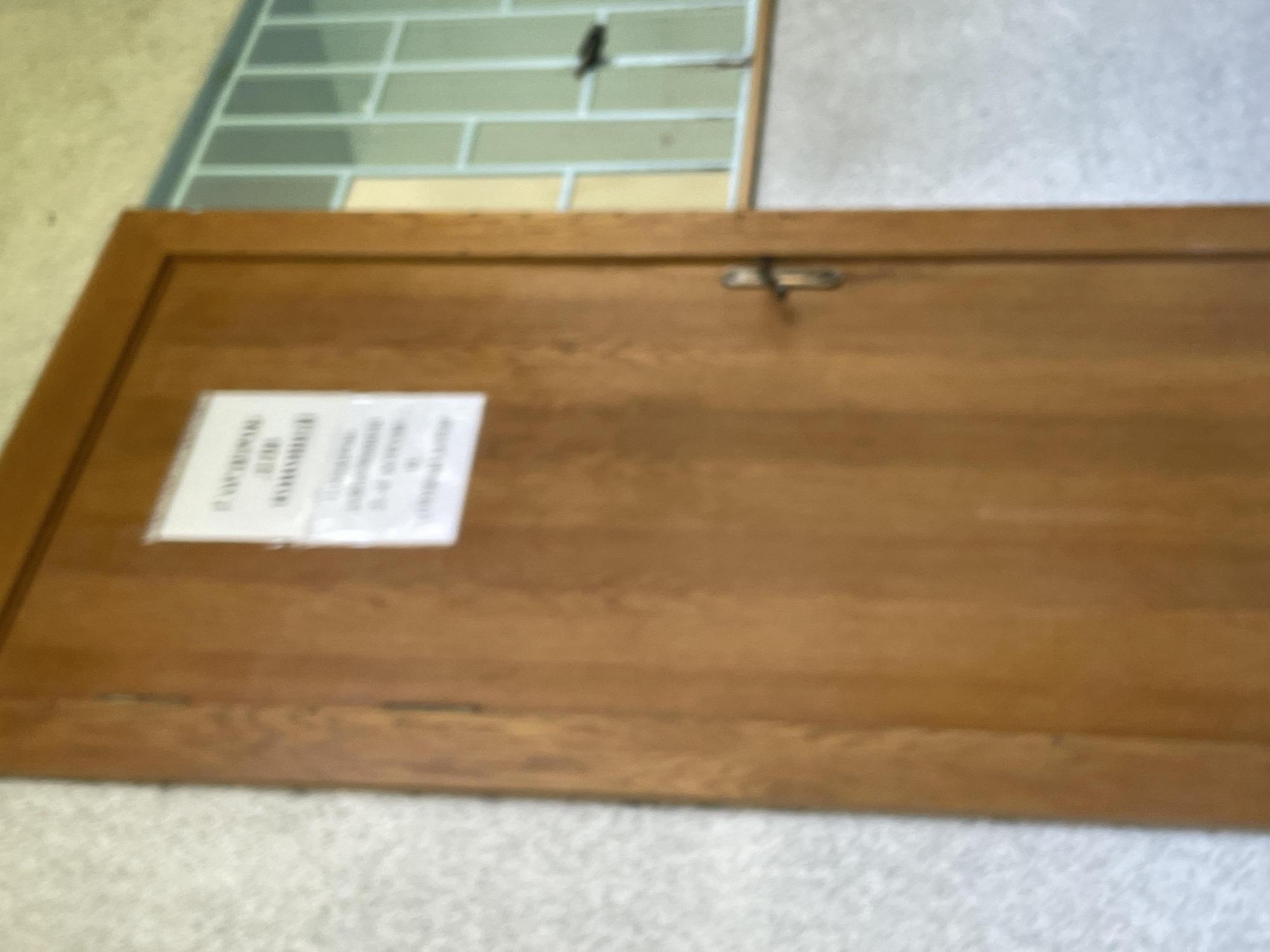 A medium shot captures a wooden door with a sign on it and a window with a blue grill to the right. The door is a light brown wood grain and has a silver handle. The sign on the door is white with black text, but the text is illegible. The window has a blue metal grill with a grid pattern and a white sill below it. The wall to the left of the door is textured and light grey. The floor is not visible. The lighting appears to be natural light coming from the window.