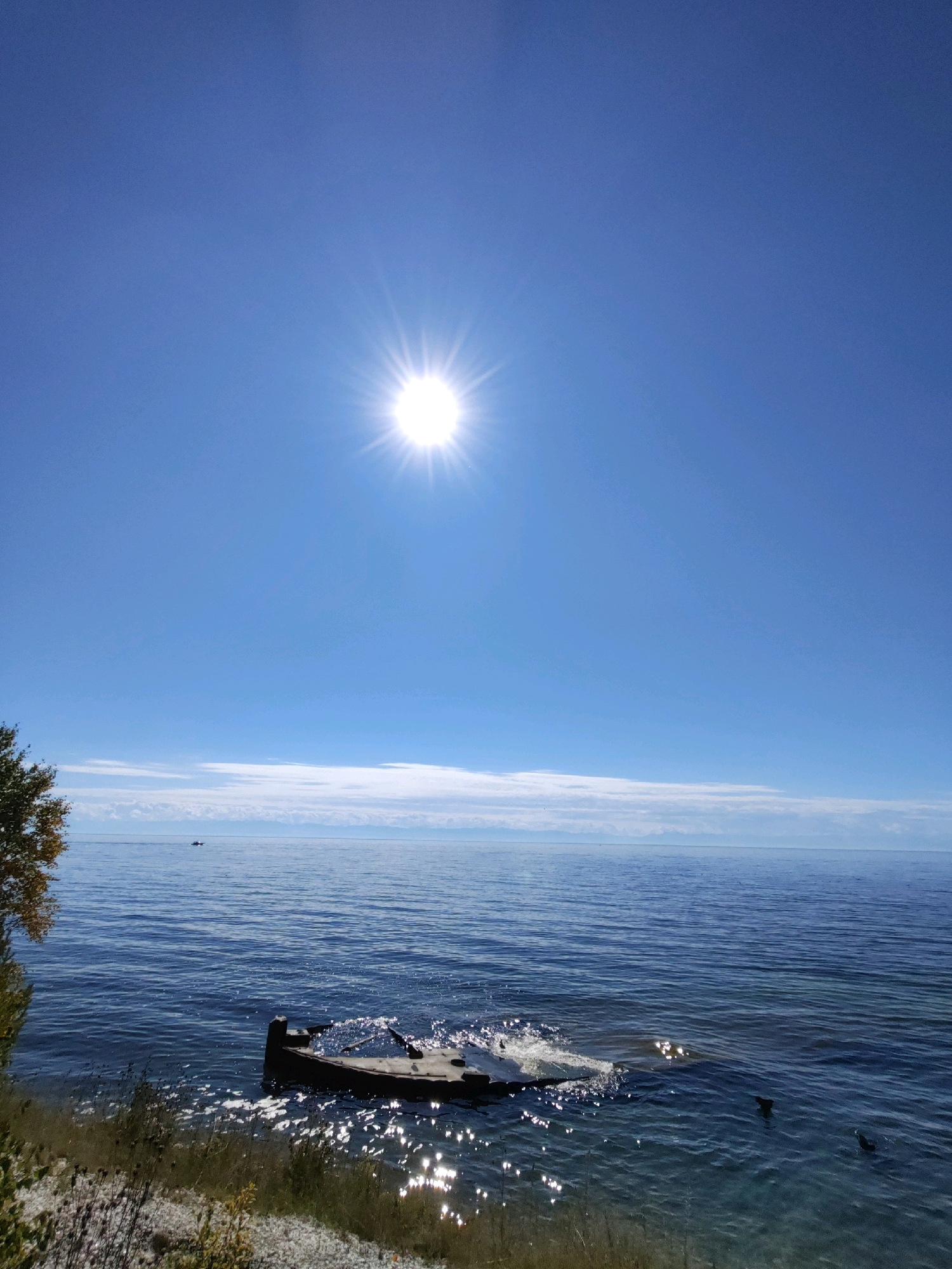 The image depicts a sunny day on the shore of Lake Baikal, Russia. The sun is prominently featured in the upper portion of the frame, radiating light. The vast blue expanse of the lake stretches towards the horizon, where a thin line of clouds is visible, suggesting a clear sky with some atmospheric haze. In the foreground, on the left side, the edge of the lake is visible with dry grass and some foliage. Partially submerged in the water, near the shore, is the wreckage of a boat, its rusty hull broken and uneven, with parts of it submerged and others exposed to the air. The water around the wreckage is choppy, with sunlight glinting off the ripples. In the distance, a small boat can be seen on the water. The overall scene conveys a sense of natural beauty and the remnants of human activity against the backdrop of a serene, expansive lake.