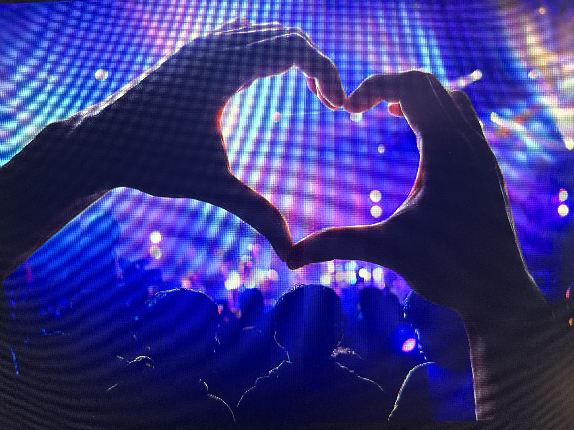 The image displays a pair of human hands positioned in the foreground, forming a heart shape through the arrangement of thumbs and forefingers. The hands are silhouetted, appearing dark against the luminous background, and exhibit noticeable pixelation across their surface, indicating the image is a photograph of a digital screen.
The background features a dynamic array of blurred, circular, and streaked artificial lights, predominantly in shades of blue, purple, with accents of white, pink, and orange. These lights are characteristic of a concert, festival, or similar large-scale event. Below the heart-forming hands, the dark silhouettes of multiple individuals' heads and shoulders are visible, representing an audience attending the event. No specific architectural features, geographic landmarks, or textual information are discernible within the image to forensically confirm its location within the city of Varna, Bulgaria. The primary subjects are the expressive hands and the vibrant, illuminated atmosphere of a large public gathering.