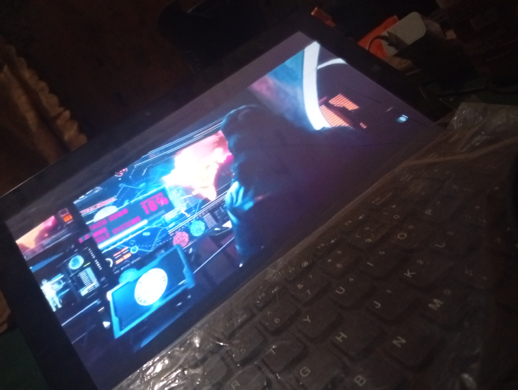The image shows a close-up, angled view of a laptop screen displaying a futuristic interface with glowing blue and pink elements, and a silhouette of a person's head and shoulders. The screen content includes what appears to be data readouts and circular graphics, with text such as "78%" clearly visible. The laptop keyboard is partially visible in the foreground, covered by a clear plastic sheet. The background is dark and appears to be an indoor setting, possibly a room, with some blurred objects and textures suggesting furniture or other items. The lighting is dim, with the primary illumination coming from the laptop screen. The location in Zongoma Kyari, Nigeria is not discernible from the visual information provided in the image.